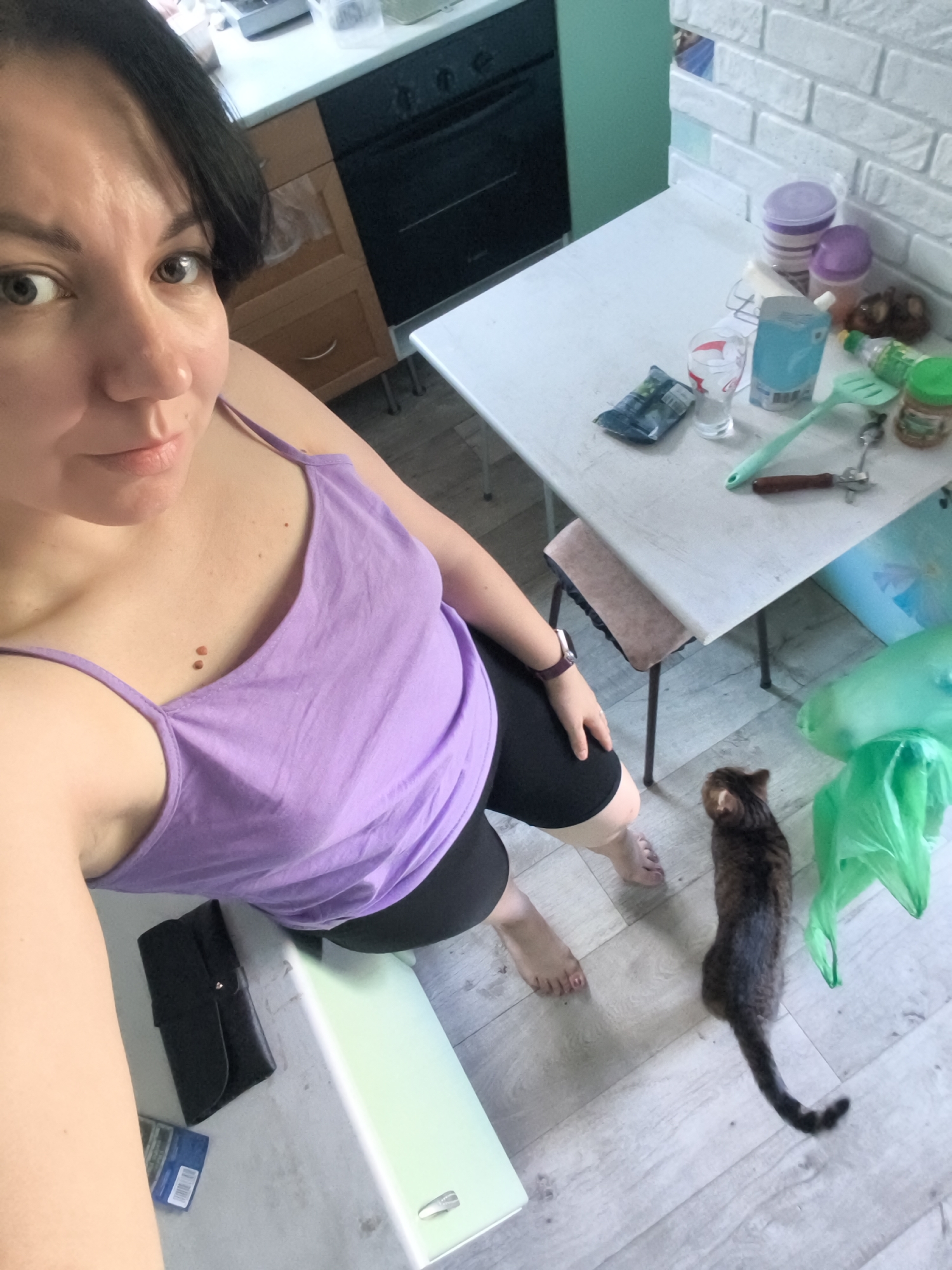 The image is a high-angle, full-body selfie taken indoors, likely in a kitchen or dining area. A woman with dark hair and fair skin is the primary subject, positioned in the lower left of the frame, looking towards the camera. She is wearing a purple tank top and black shorts. Her left hand is resting on her knee, and she is barefoot.
To her right, a small, tabby cat with dark fur is walking away from the camera on a light-colored, wood-plank floor.
In the background, a white-topped table is cluttered with various items, including containers, a glass, a packet, a green utensil, and a bottle. Behind the table, a dark oven and wooden cabinets are visible. The walls appear to be white brick in some sections and a light blue in others. The overall environment suggests a residential indoor setting. The specific location in Orenburg, Russia cannot be determined from the image alone.