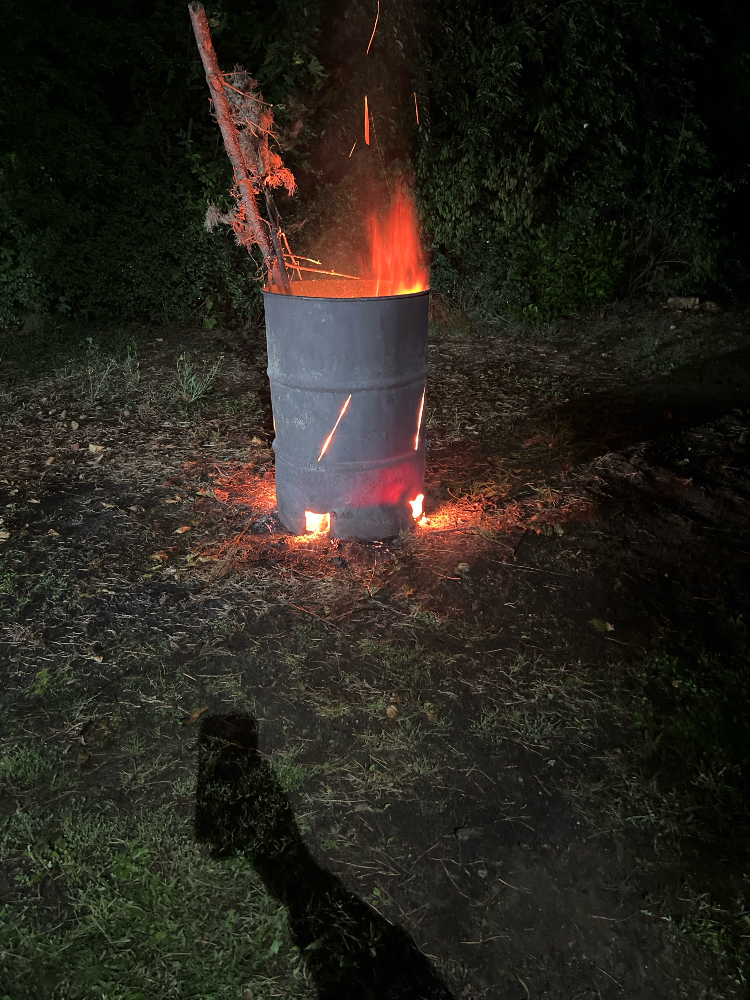 A barrel is burning in a grassy area at night. The barrel is dark gray metal and has a piece of wood sticking out of it with some branches. Flames are visible inside and coming out of the top and some openings near the bottom of the barrel. There are also embers around the base of the barrel. The ground is covered in grass, dirt, and fallen leaves. In the background, there are dark green trees and bushes. A shadow of a person is visible on the ground in the foreground.