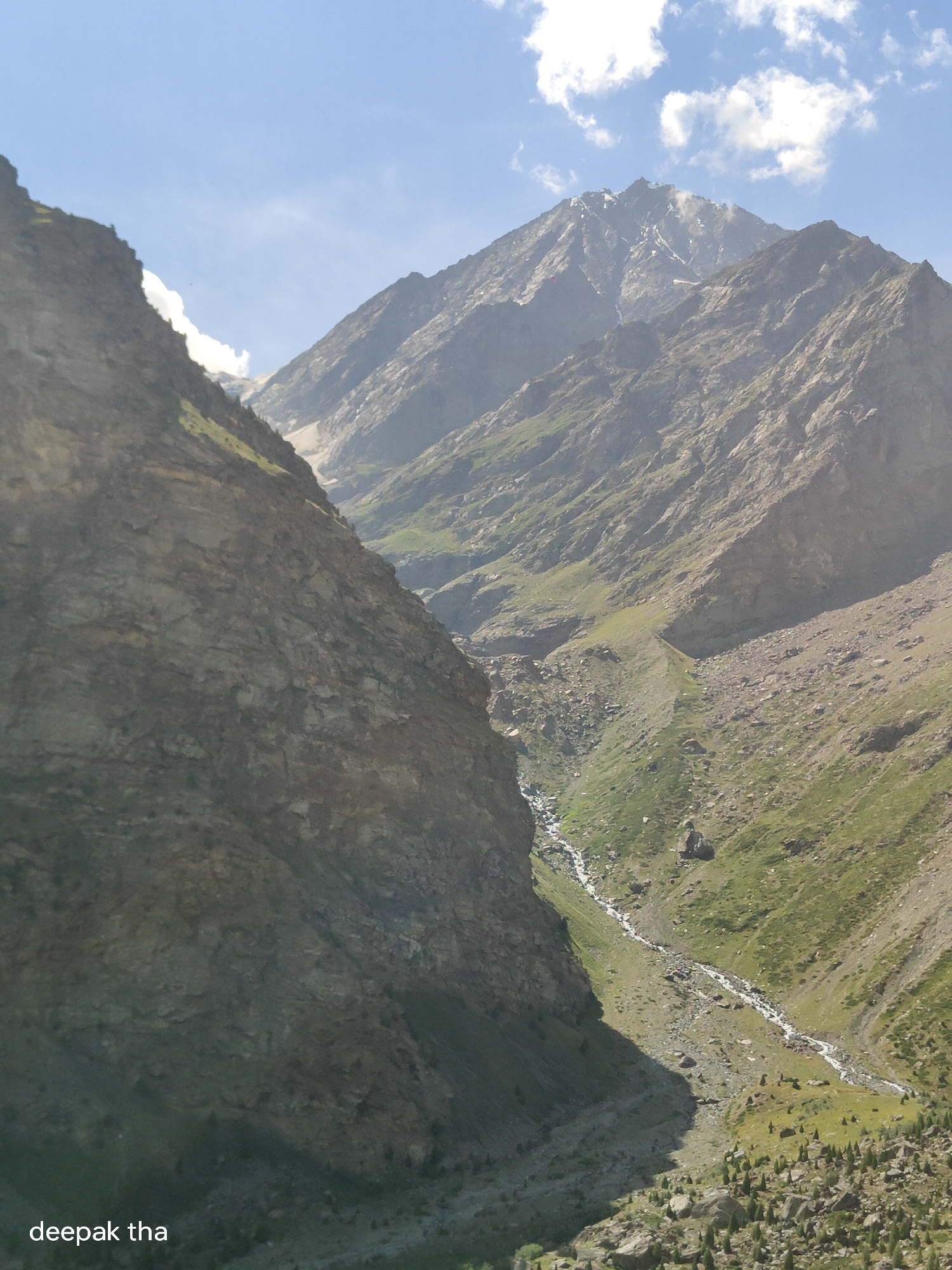 The image shows a mountain landscape with a narrow valley. On the left, a steep, rocky cliff face dominates the foreground. A smaller, more gradual slope covered in green vegetation and scattered rocks descends into the valley. A thin, winding stream flows down the center of the valley. In the background, more rugged, rocky mountain peaks rise up, with some patches of snow visible near the summits. The sky is blue with scattered white clouds. The overall scene is a natural, rugged mountain environment. The text "deepak tha" is visible in the lower left corner of the image. There are no people visible in the image.