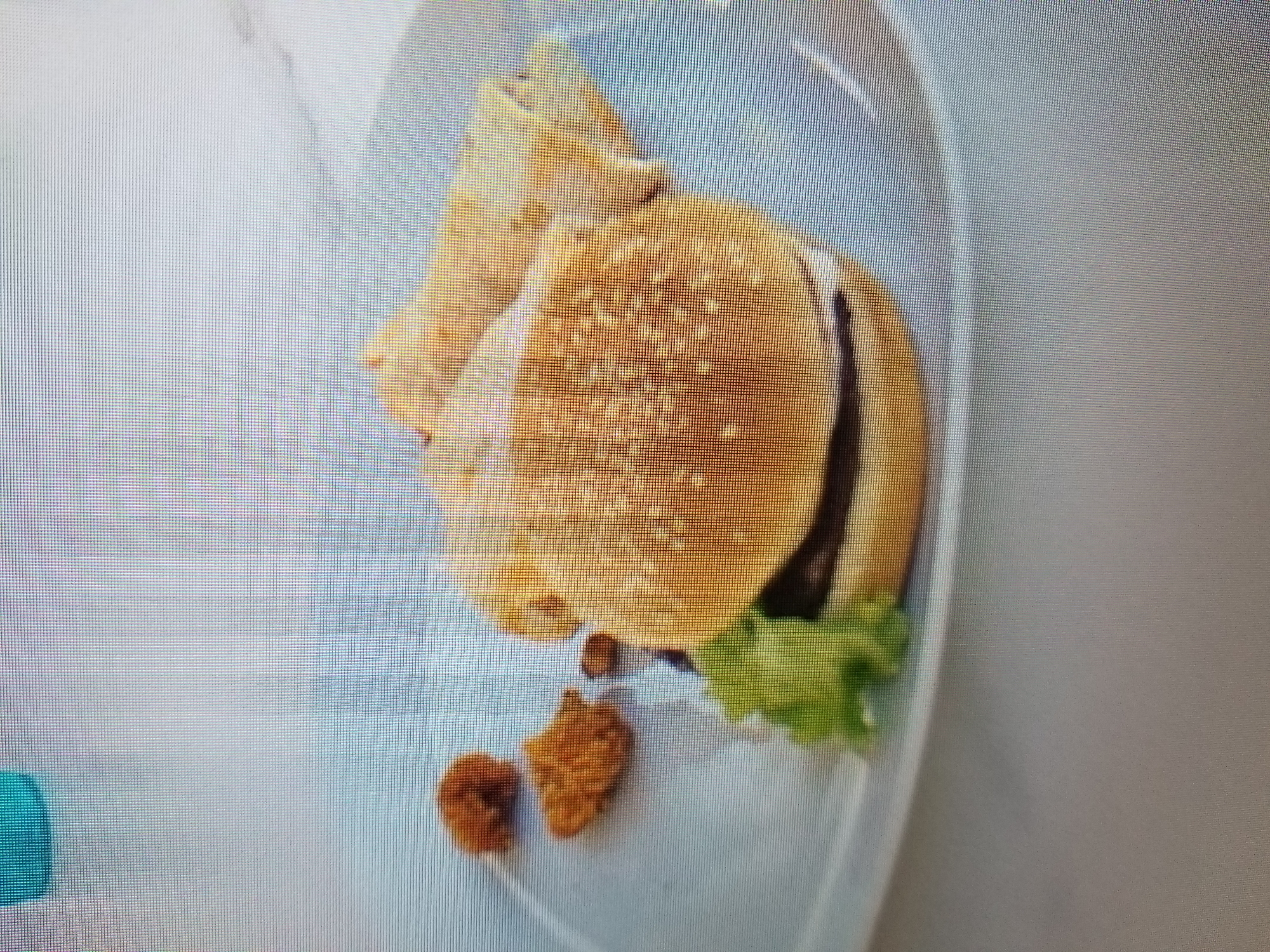 A close-up, slightly blurry, eye-level, medium shot shows a white square plate with food. On the plate is a hamburger, two or three pieces of fried chicken, and some rolled-up food items. The hamburger has a sesame seed bun, a dark patty, and a piece of green lettuce. The fried chicken pieces are golden brown and irregular in shape. The rolled-up items appear to be burritos or wraps, with a light-colored filling. The plate is on a light-colored, smooth surface, possibly a table. A small portion of a blue object is visible in the upper left corner of the frame. The image is too low-resolution to provide specific details about the location in Tiraspol, Moldova.