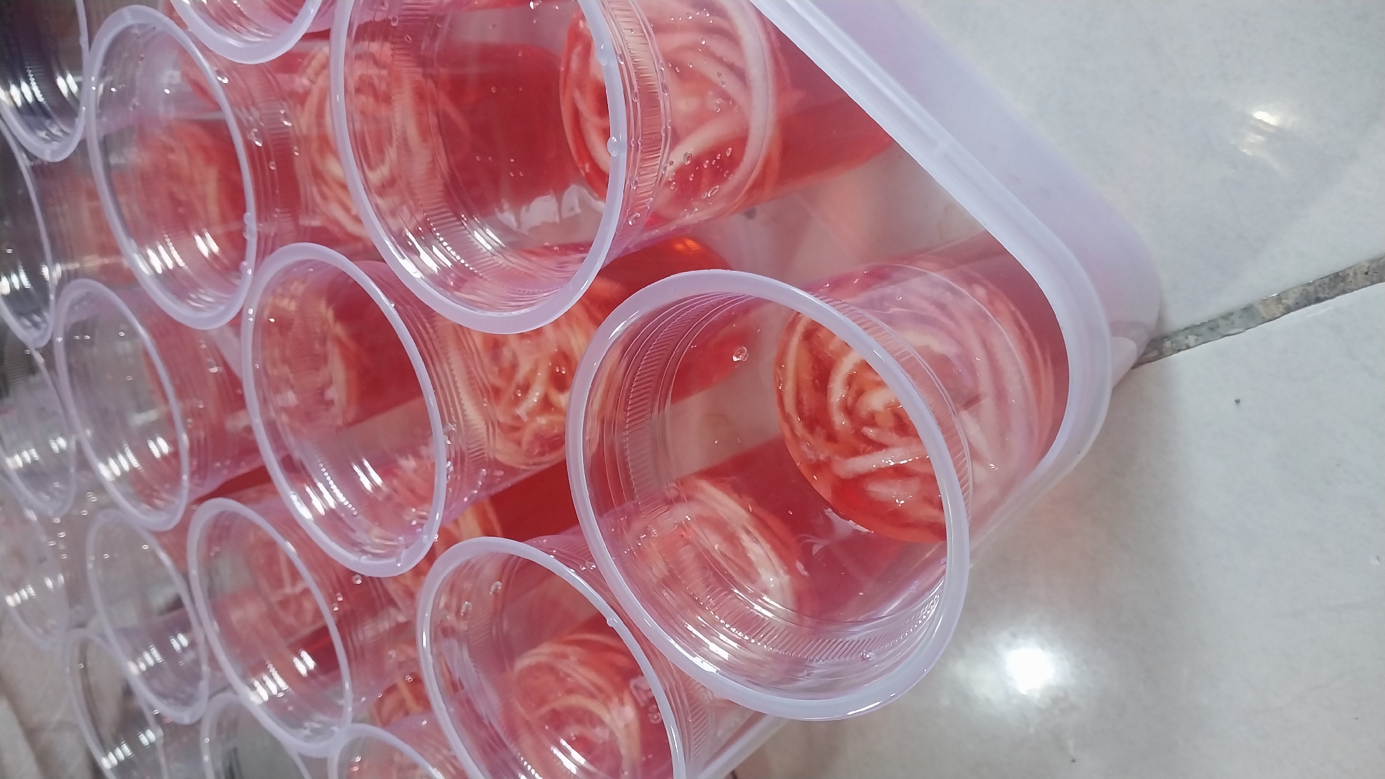 The image displays a tray filled with numerous clear plastic cups, each containing a pink liquid with a swirl of white and pink substance. The cups are arranged in rows and appear to be a dessert, possibly jelly or pudding, with a layered or swirled texture. The tray itself is a light pink plastic container. The cups and tray are placed on a tiled surface that is light gray with visible grout lines. The overall scene is domestic and appears to be indoors. There are no people visible in the image, and the lighting suggests it could be daytime or a well-lit indoor space.
