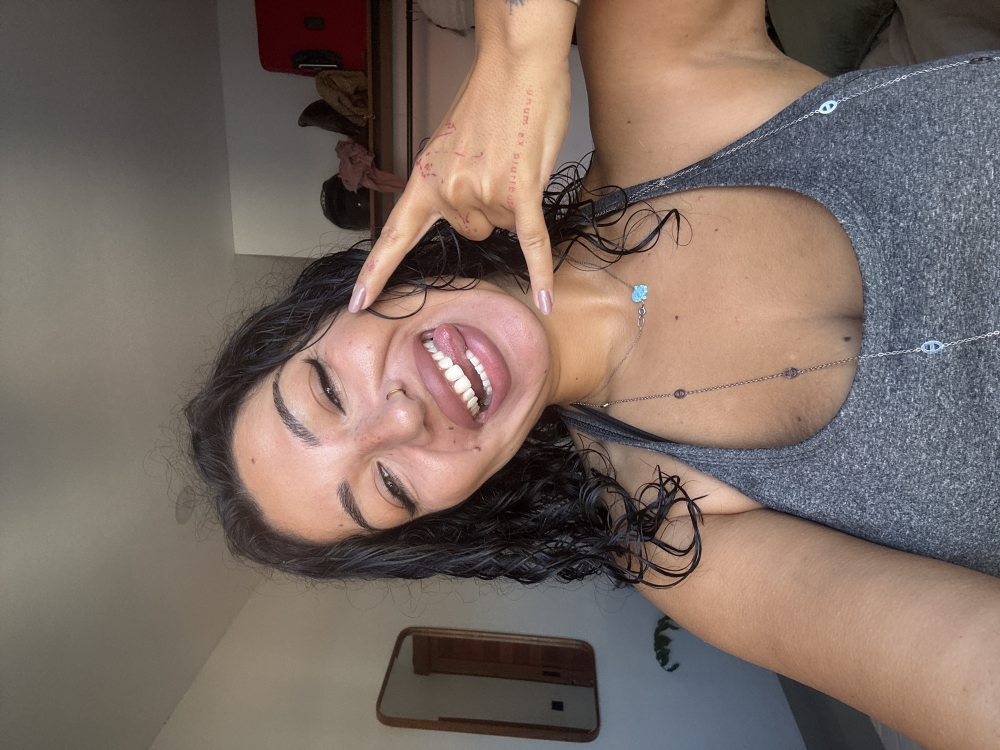 A young woman with dark, wet hair, dark eyebrows, and light brown eyes is posing for a selfie indoors. She is wearing a grey tank top and multiple necklaces. Her tongue is sticking out, and she is holding up her right hand with two fingers extended, forming a "rock on" gesture. She has light-colored nail polish on her fingernails. There are faint red markings on the back of her hand, possibly tattoos or temporary ink. A mirror is visible on the left side of the frame, reflecting part of a room. In the background, on a shelf, are several items including a red suitcase, some clothing items, and a dark helmet. The lighting suggests it is daytime, with light streaming in from an unseen window. The woman's skin has freckles and a beauty mark.