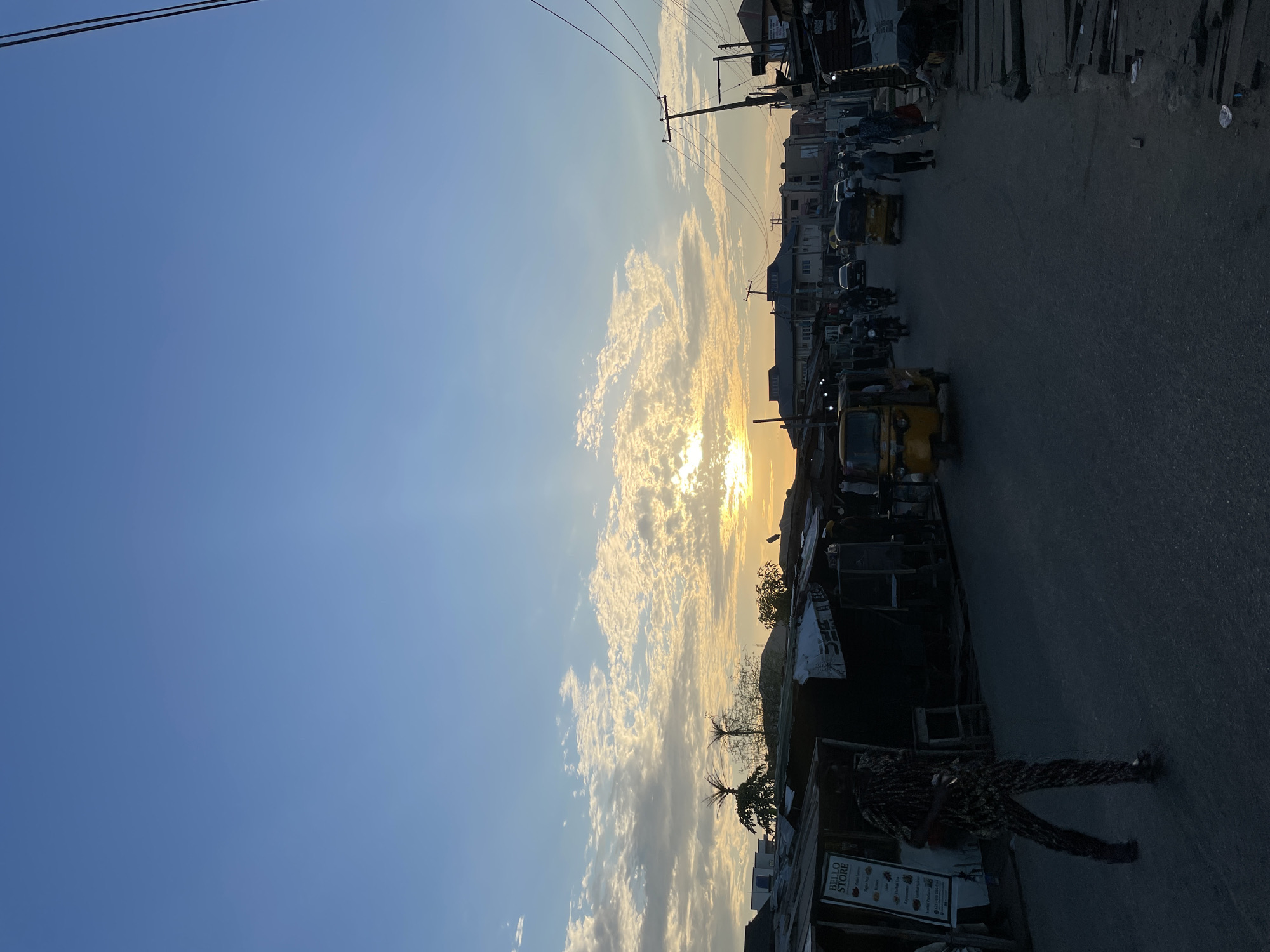 The image shows a street scene in Isheri, Nigeria, at what appears to be dusk or dawn. The sky is visible with white clouds as the sun sits low on the horizon. The road stretches into the distance with buildings and power lines visible on the right. There are vehicles in the street, including small commercial auto rickshaws (tuk-tuks) and potentially cars and motorcycles. There are also people on the street, and one person is particularly visible in the foreground, along with storefronts and signage. The lighting suggests it is either sunrise or sunset, with silhouettes becoming pronounced.