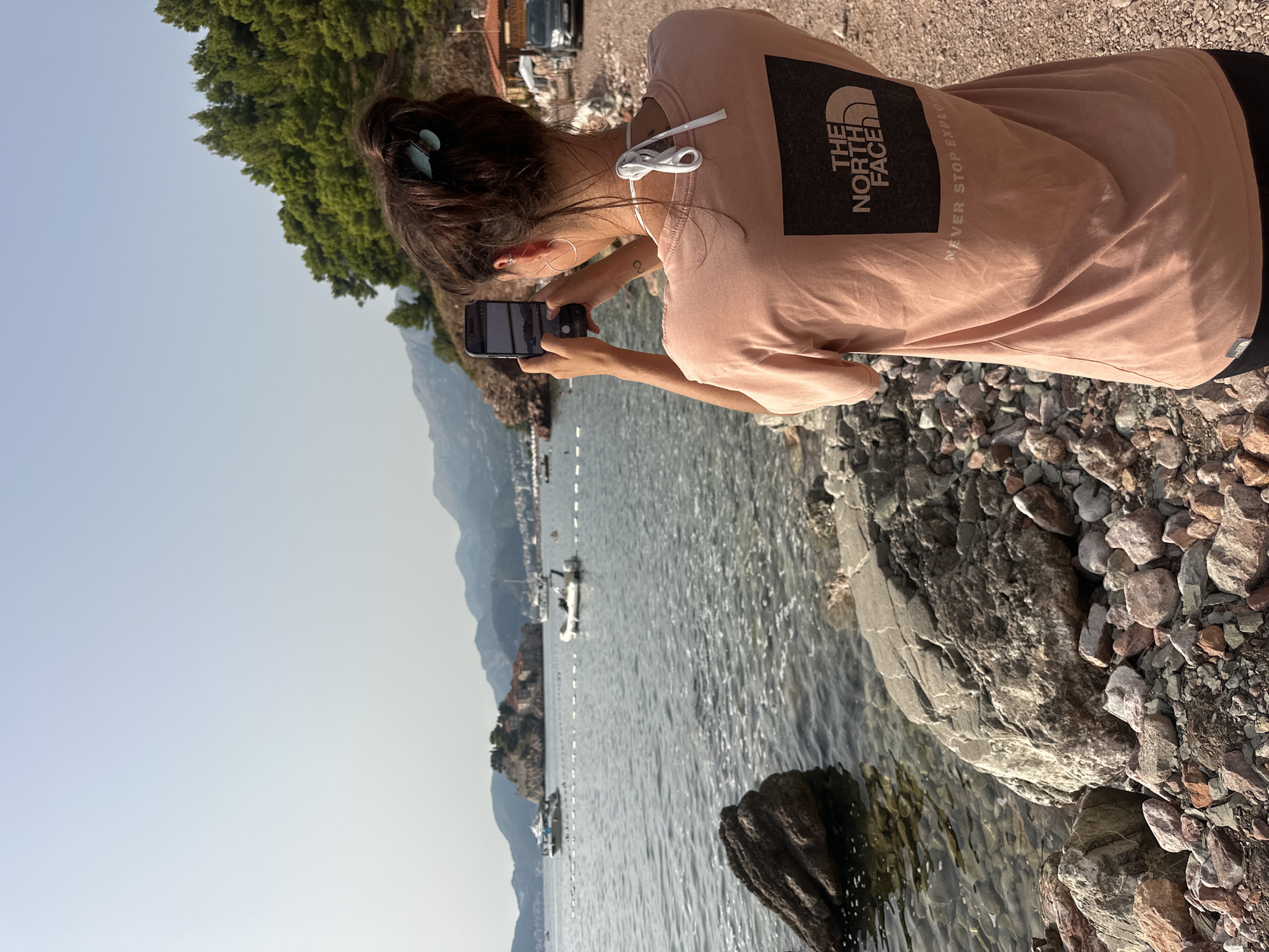 In Sveti Stefan, Montenegro, a young woman stands on a rocky shore, facing away from the camera and towards the Adriatic Sea. She is wearing a pale peach t-shirt with "The North Face" logo on the back and black shorts. She is holding a smartphone in her right hand, using it to take a photograph of the scenic view, which includes the iconic island of Sveti Stefan in the distance, dotted with buildings and surrounded by water. Several boats are anchored or moving in the bay, and a bucolic chain of boats marks a swimming area. The background features lush green trees and distant mountains, with a clear, pale blue sky overhead. The foreground is dominated by the rough texture of the rocky beach, with larger boulders and smaller pebbles.