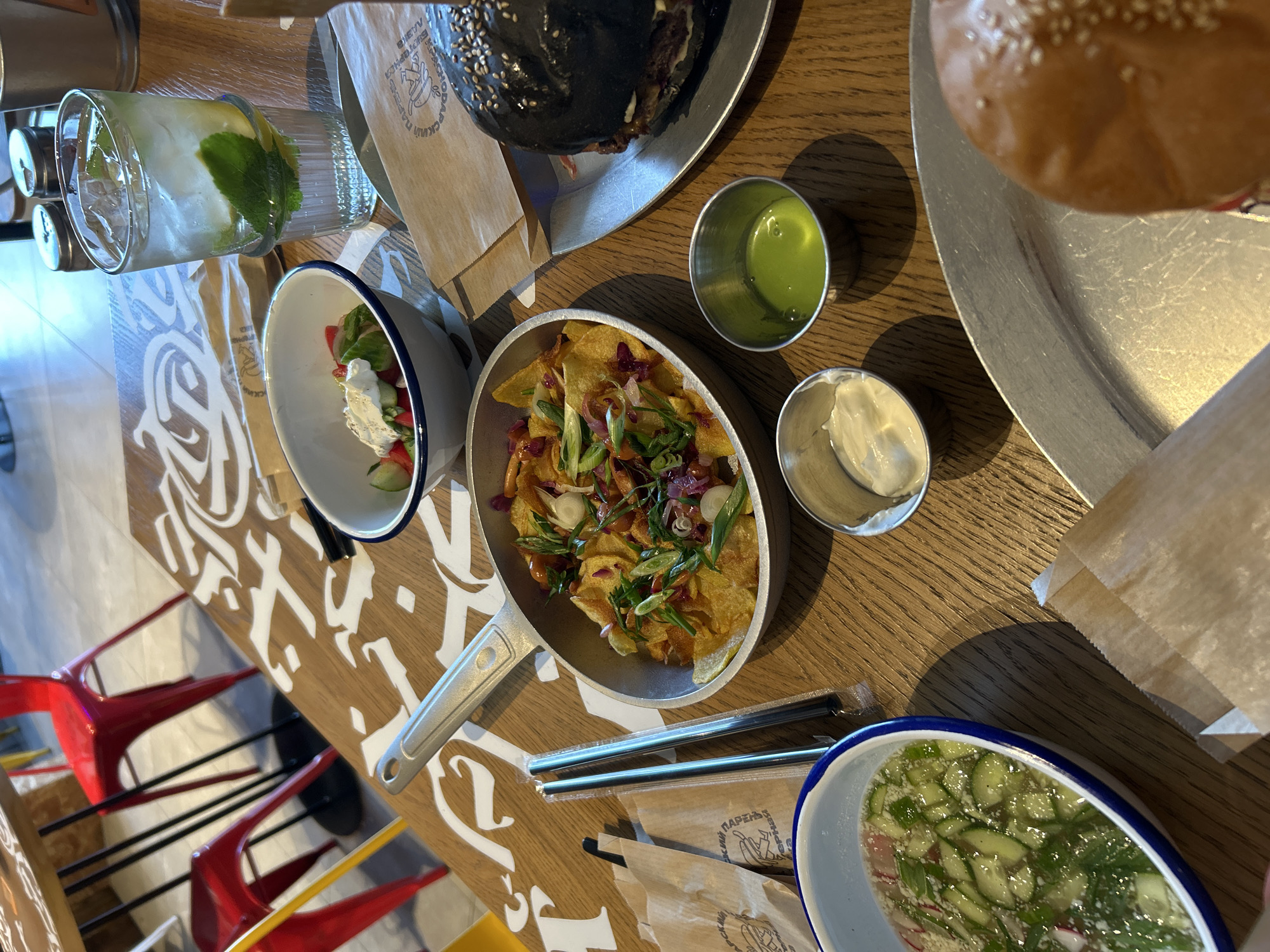 An overhead shot captures a table at a restaurant with various dishes and drinks. In the foreground, a small bowl contains a cucumber salad with radishes and herbs. To its right, two small metal ramekins hold creamy white sauce and a bright green sauce. A skillet filled with potato chips topped with scallions and red onions sits on the table. A black burger with sesame seeds on the bun is on a metal plate to the right of the skillet. A tall glass with a beverage, ice, mint, and lemon slices rests near the salad. Two black metal straws are bundled together in plastic wrap. In the background, red chairs are visible around a table with decorative white calligraphy. The setting appears to be an indoor dining area with wooden tables and flooring.