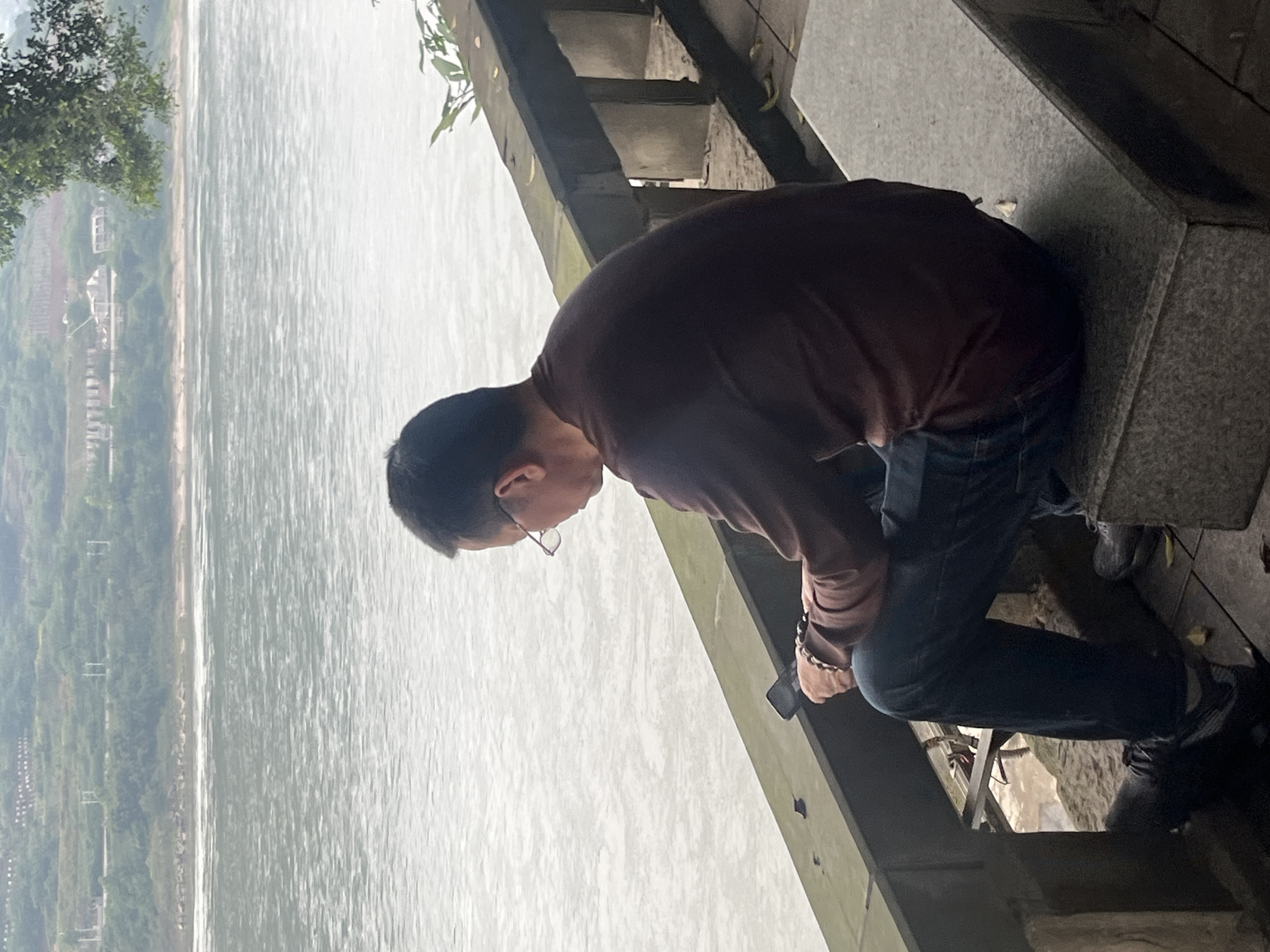 A man wearing glasses, a long-sleeved brown shirt, and blue jeans stands on stone steps in Jiangjin, China. He holds a phone in his hand and looks down towards the river. The river is wide and stretches to the horizon, with green trees and buildings lining the opposite bank.