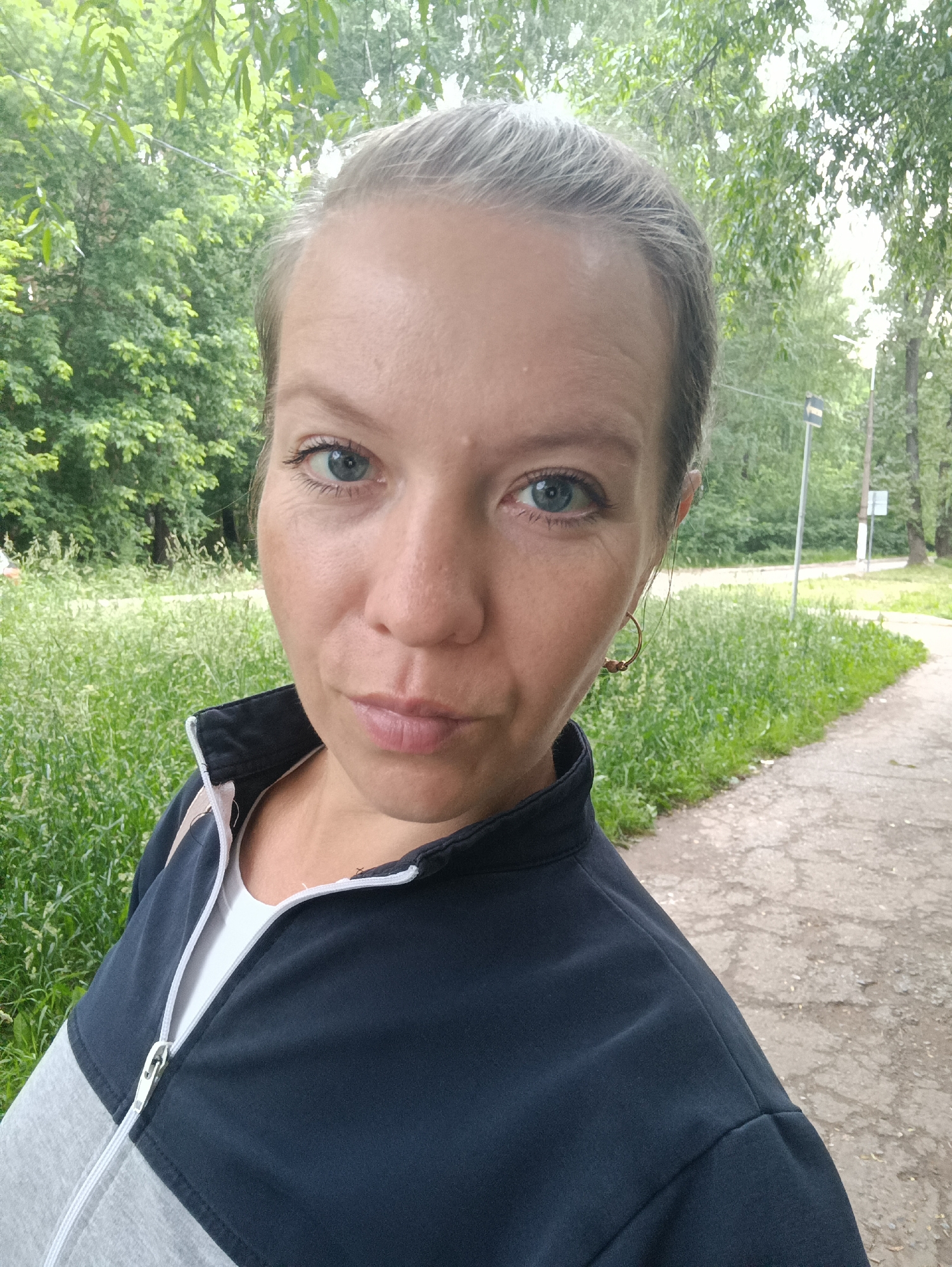 An outdoor, close-up, head-and-shoulders selfie of a woman with blonde hair pulled back in a ponytail. She is wearing a dark blue and gray zip-up jacket over a white t-shirt and a single hoop earring on her right ear. Her eyes are blue and she is looking at the camera with a neutral expression. The background is slightly blurred and consists of green trees and grass, and a cracked asphalt path. A street sign is visible in the far background. The image was taken on a cloudy day in the city of Chaikovsky, Russia.