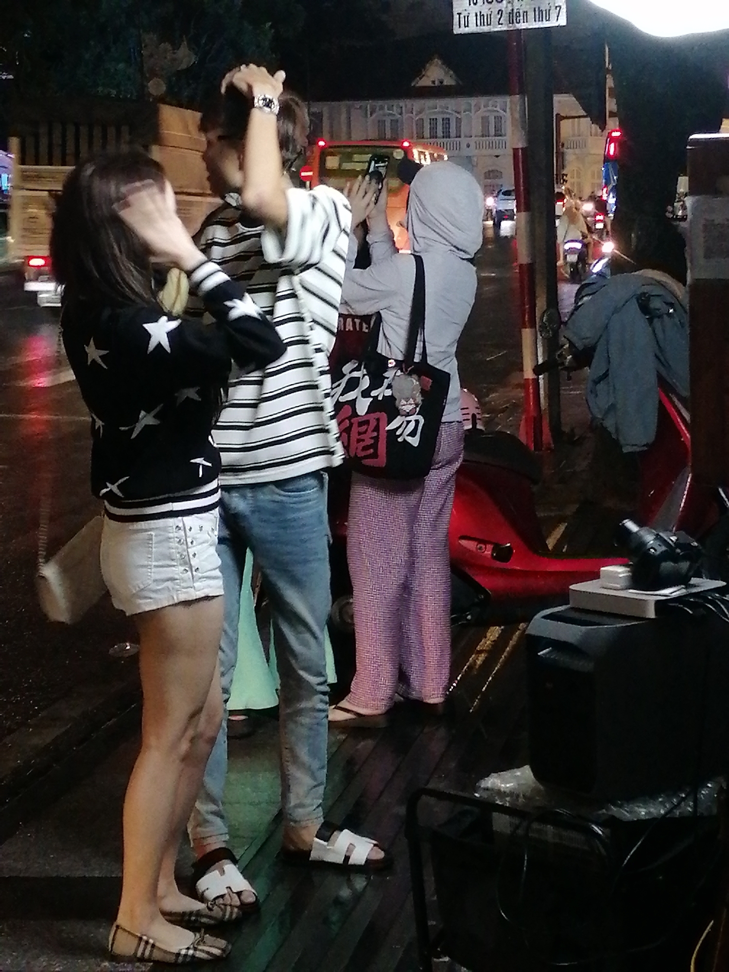 The image shows a street scene at night in Da Lat, Vietnam. In the foreground, three individuals are present. On the left, a woman with dark hair is wearing a black jacket with white stars and white studded shorts. She is holding her hand to her face. Next to her, a man with dark hair is wearing a striped t-shirt and jeans. He is looking at his phone. To the right, another person is wearing a grey hooded jacket and patterned pants, holding a black bag with a cartoon character. They are also holding a phone. Several motorcycles are visible in the background, as well as some buildings and street lights. The ground appears to be wet, suggesting recent rain.