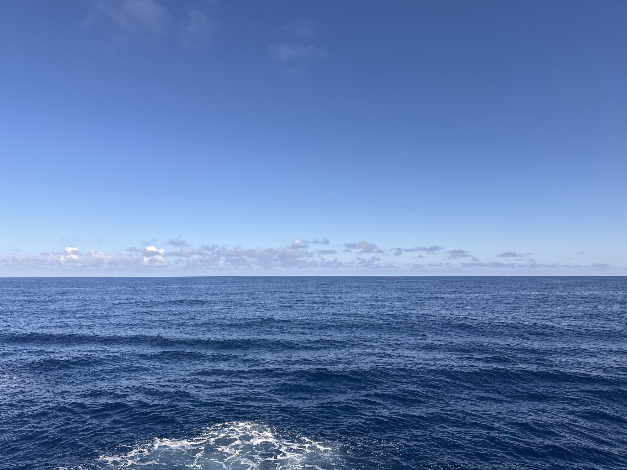 The image shows a calm sea under a clear, blue sky with a few scattered clouds. The water is a deep blue color, with gentle waves creating subtle patterns on the surface. White foam is visible in the foreground, likely from the wake of a boat. The horizon line is distinct but blurred by the clouds. The location is the open ocean.