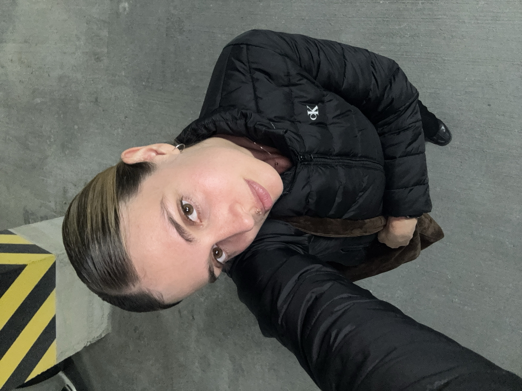 The image is a selfie of a person, likely female, wearing a black Calvin Klein puffer jacket. The person has light brown eyes and light brown hair slicked back. They have a piercing on their upper lip and are wearing an earring. The location appears to be a parking garage, as indicated by the gray concrete floor and the black and yellow hazard marker in the background. The person is holding the camera at an upward angle, capturing their face and the upper part of their body. They are also holding a brown bag.