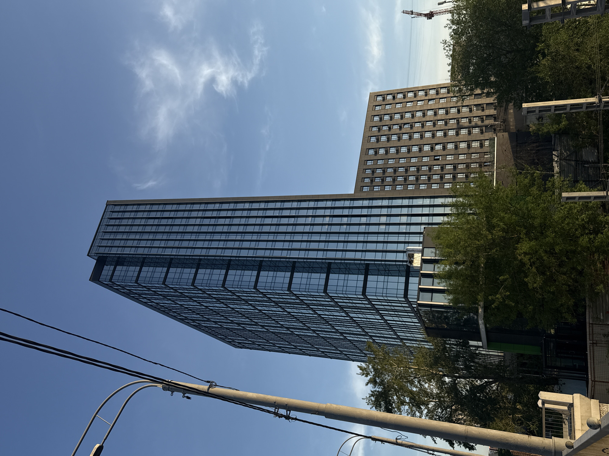The image shows two tall buildings under a blue sky with scattered clouds. The taller building, located on the left, is a modern glass skyscraper, reflecting the sky in its windows. To the right, there is a second, more traditional building with a gray façade and regularly spaced windows. Trees partially obscure the base of both buildings. In the foreground, there are utility poles and power lines stretching across the frame. In the background, a construction crane is visible. The location appears to be in an urban environment, specifically Moscow, Russia, based on the architectural style and the general cityscape.