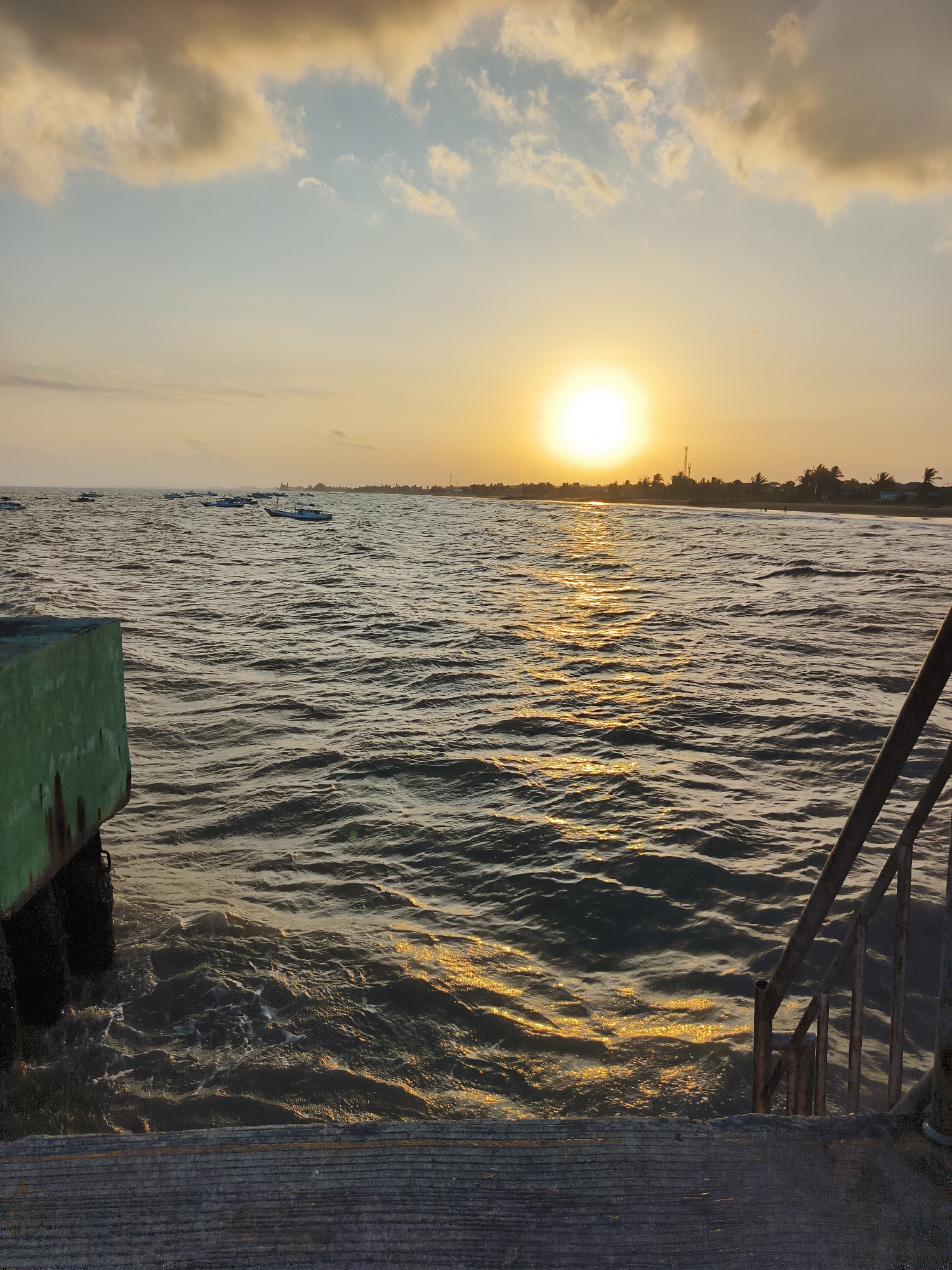 At the city of Kupang Berkah Jaya, Indonesia, a serene sunset casts a warm, golden glow over the rippling ocean. The sun, a bright orb, hangs low in the sky, partially obscured by wispy clouds tinged with orange and yellow. Its reflection shimmers on the water's surface, creating a path of light that stretches towards the horizon.
Several small boats are scattered across the expanse of the sea, their silhouettes dark against the illuminated water. In the distance, a coastline is visible, lined with palm trees and a few buildings, suggesting a coastal town. The foreground features a concrete structure with a green, weathered surface, likely part of a pier or dock, on the left. On the right, a rusted metal staircase descends towards the water, its railings adding a rustic texture to the scene. The waves are gentle, creating a calm and peaceful atmosphere.