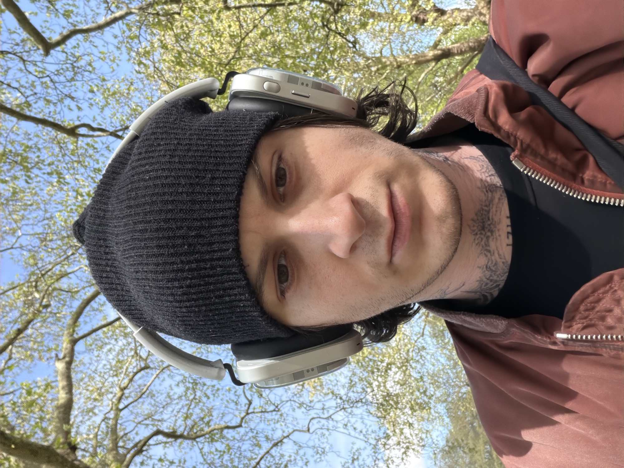 The rotated image captures a close-up, low-angle perspective of a person outdoors, likely in London, United Kingdom. The subject, who appears to be male, looks directly at the camera with a neutral expression. They wear a dark, possibly black, knitted beanie and light-colored over-ear headphones, which rest partially on the beanie and partially on their dark hair. Tattoos are visible on the left side of their neck, extending under their jawline.
The person is dressed in a reddish-brown jacket with a visible silver zipper, worn over a dark shirt. The background features a bright blue sky with scattered white clouds, seen through the branches of trees with lush, light green leaves, indicative of spring or early summer. The sunlight illuminates the scene, suggesting a clear and pleasant daytime. The person seems to be engaging in a casual outdoor activity, possibly listening to music or a podcast while enjoying a walk or relaxing in a park. There is no visible text in the image.