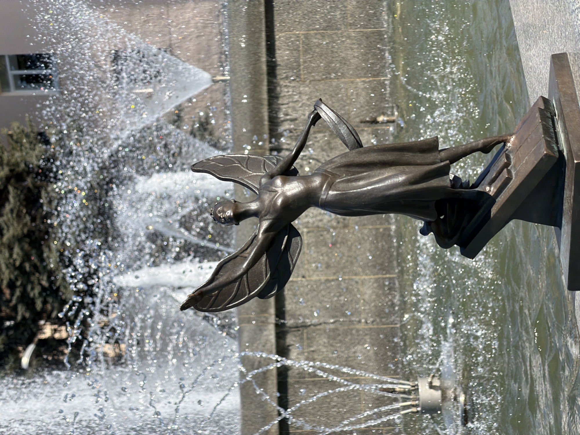 A sculpture of a female figure with wings is prominently displayed. The figure appears to be made of dark metal and has a slightly weathered appearance. It is positioned on a pedestal within a fountain, surrounded by multiple water jets. The water creates a dynamic effect of cascading droplets. The fountain is located in the city of Chaikovsky, Russia. In the background, parts of buildings and greenery are visible, suggesting an urban setting. The photograph is taken at an angle.