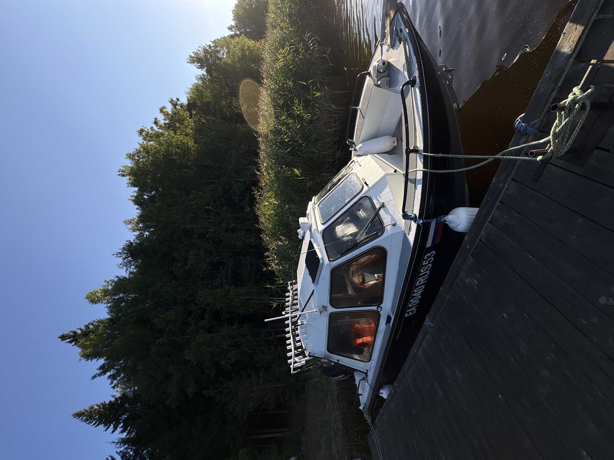 In Rautalakhti, Russia, a white and black motorboat is docked at a wooden pier. The boat has a cabin with windows and an orange life vest is visible inside. A fishing rod holder is mounted on the roof of the cabin. The boat's registration number, "EA0040 RUS53," is visible on the hull. The water is calm, and the pier is made of dark wood planks. A dense line of green trees forms the background, with a clear blue sky above. The sun is shining, casting a warm light on the scene.