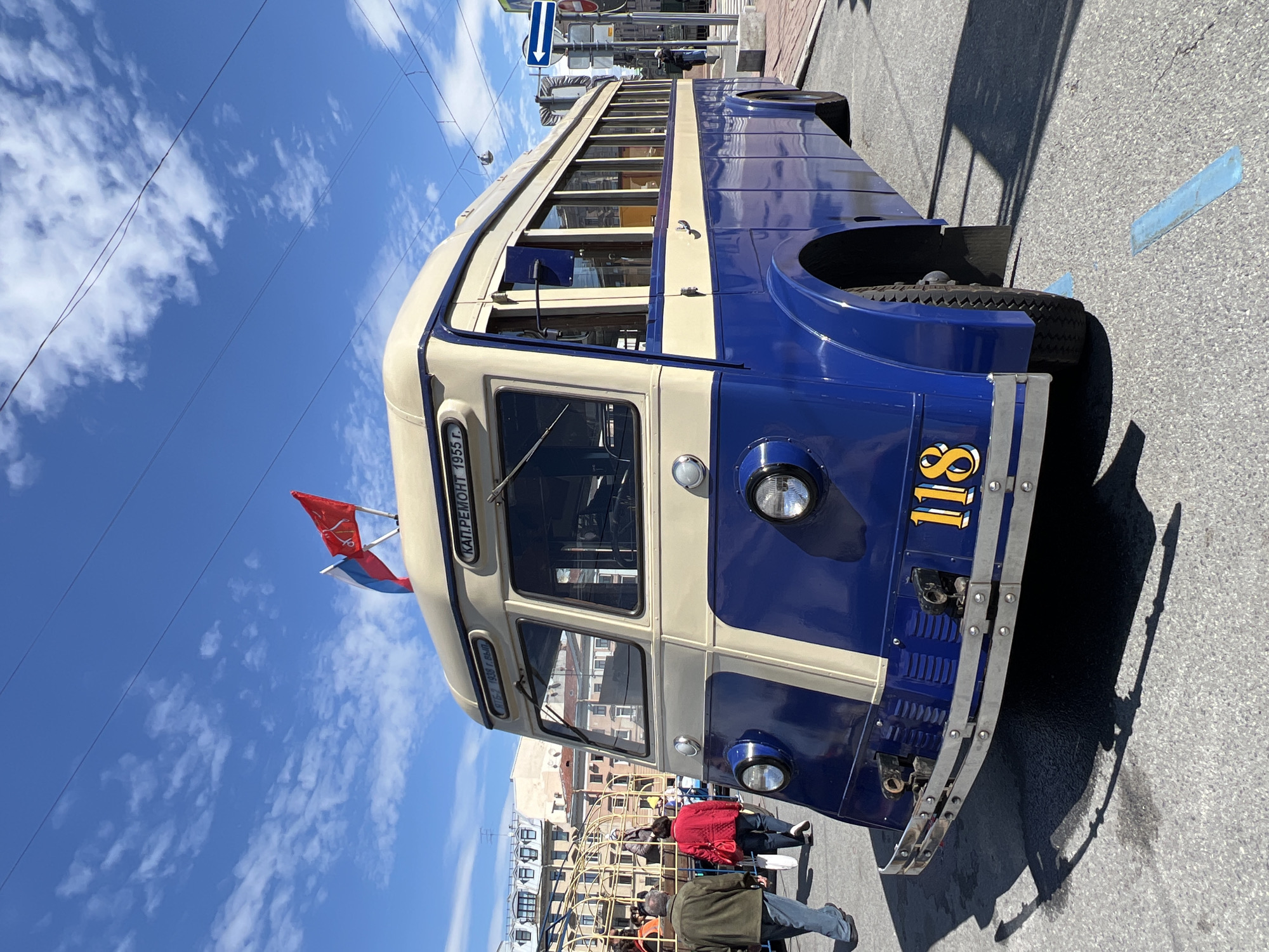 A vintage blue and cream trolleybus, likely a restored model, is parked on a city street on a sunny day. The number "118" is prominently displayed in yellow on the front of the trolleybus. A small Russian flag and another red flag with a logo are attached near the top front corner. People are walking near a playground structure in the background. The location is the city of Saint Petersburg, Russia.