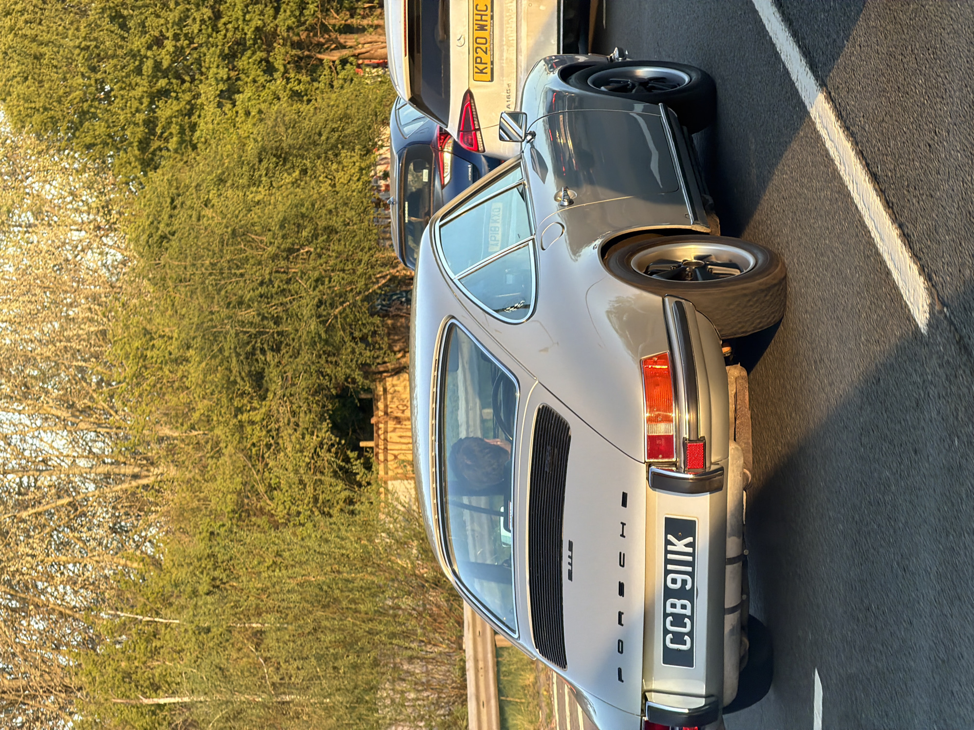 The image depicts a light grey Porsche 911 on a road, likely a motorway, given the presence of white lane markings. The car's license plate reads "CCB 911K." Behind the Porsche, partially visible, are two other vehicles, a dark blue car, and a light-colored car with the license plate "KP20 WHC." The surrounding environment includes trees along the roadside, suggesting a suburban or rural setting. It's not possible to confirm the exact location in London or the United Kingdom based on the visual information available.