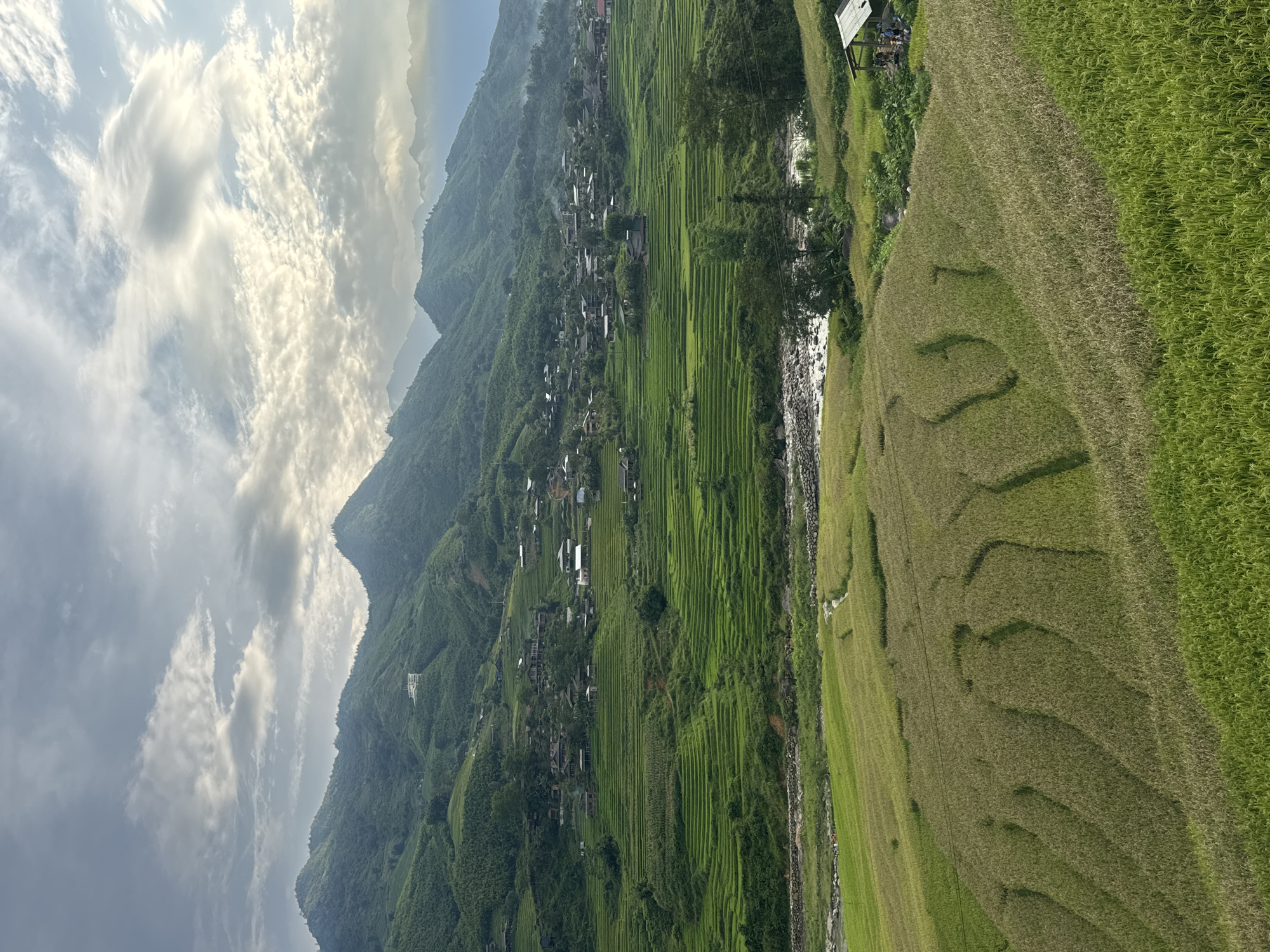 This is a landscape photograph of terraced rice fields in Tả Van, Vietnam. In the foreground, there are fields of partially harvested rice, with some areas of green rice stalks and other areas of golden-brown rice ready for harvest. The terraces are sculpted into the rolling hills, creating a natural staircase effect.
In the middle ground, a narrow, rocky river flows through the valley. On the right side of the river, a small, rustic wooden structure with a tin roof stands next to a group of people. The people appear to be gathered near the structure, possibly taking a break or observing the scenery. Further back, more vibrant green rice terraces climb the slopes of the surrounding mountains. Scattered throughout these terraces are small villages with traditional houses, characterized by their dark roofs.
The background is dominated by lush green mountains that recede into the distance, with layers of peaks creating a sense of depth. The sky above is filled with a dramatic display of clouds, with patches of sunlight breaking through, casting a soft, ethereal light on the landscape. The overall impression is one of serene natural beauty and rural life in Vietnam.