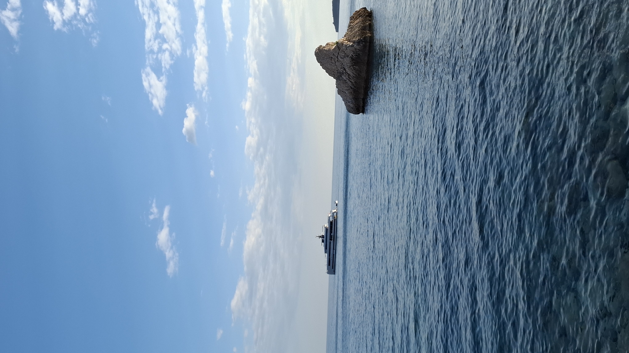 A sideways shot depicts a scene near Budva, Montenegro. The sky is blue with scattered white clouds on the left side. The ocean fills the right side of the frame. A large rock formation sits in the sea closer to the top edge of the image. Further away, a yacht is sailing on the water.