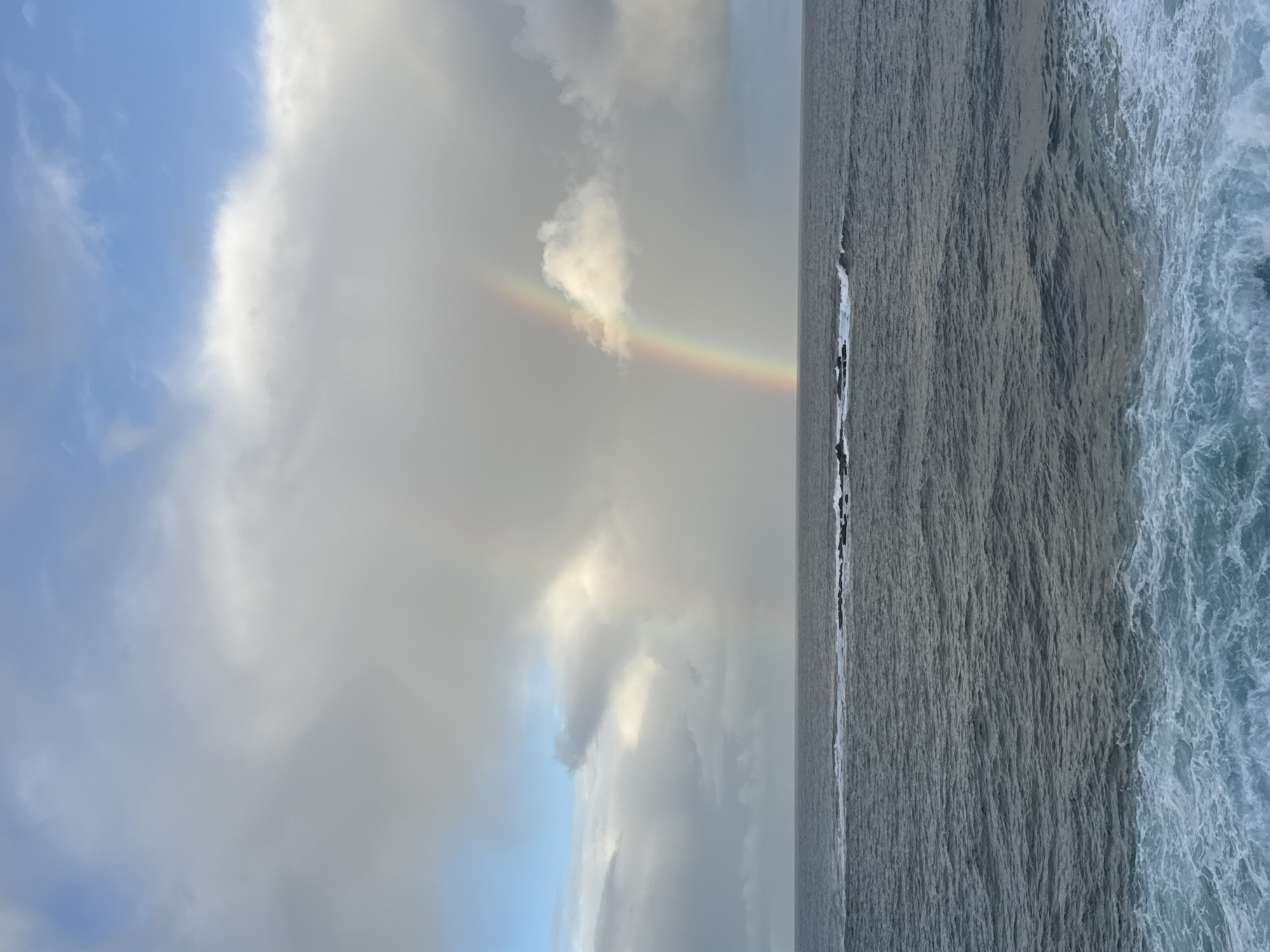 A dramatic sky with a rainbow arc is captured over a dark, choppy ocean. The rainbow, a vibrant spectrum of colors, is partially obscured by soft, gray clouds. Below, the water displays a range of dark blues and grays, with white foam cresting the waves in the foreground. A low, rocky outcrop is visible in the middle distance, breaking the surface of the water. The overall scene conveys a sense of natural power and beauty. The location is identified as Clovelly, Australia, implying an outdoor coastal environment.