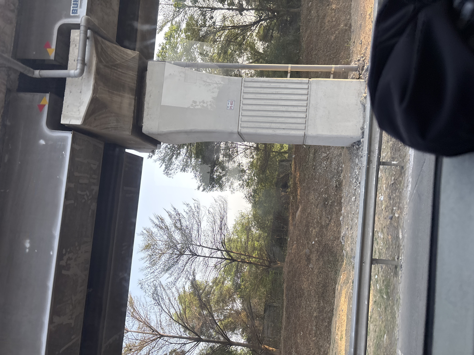 The image shows a concrete overpass support pillar in an outdoor environment. The pillar is textured with vertical ridges on its lower section and has a label that reads "PS 482". Above the pillar, the overpass structure is visible, with a gray beam supported by the pillar. There are red and yellow triangular markers on the underside of the overpass beam. The surrounding area is filled with trees and dry vegetation, with a guardrail visible in the foreground. A hint of a black garment is present in the bottom right corner. The scene appears to be captured from a moving vehicle, possibly indicating a roadside location in Jayamukti, Indonesia, given the vegetation and construction style.