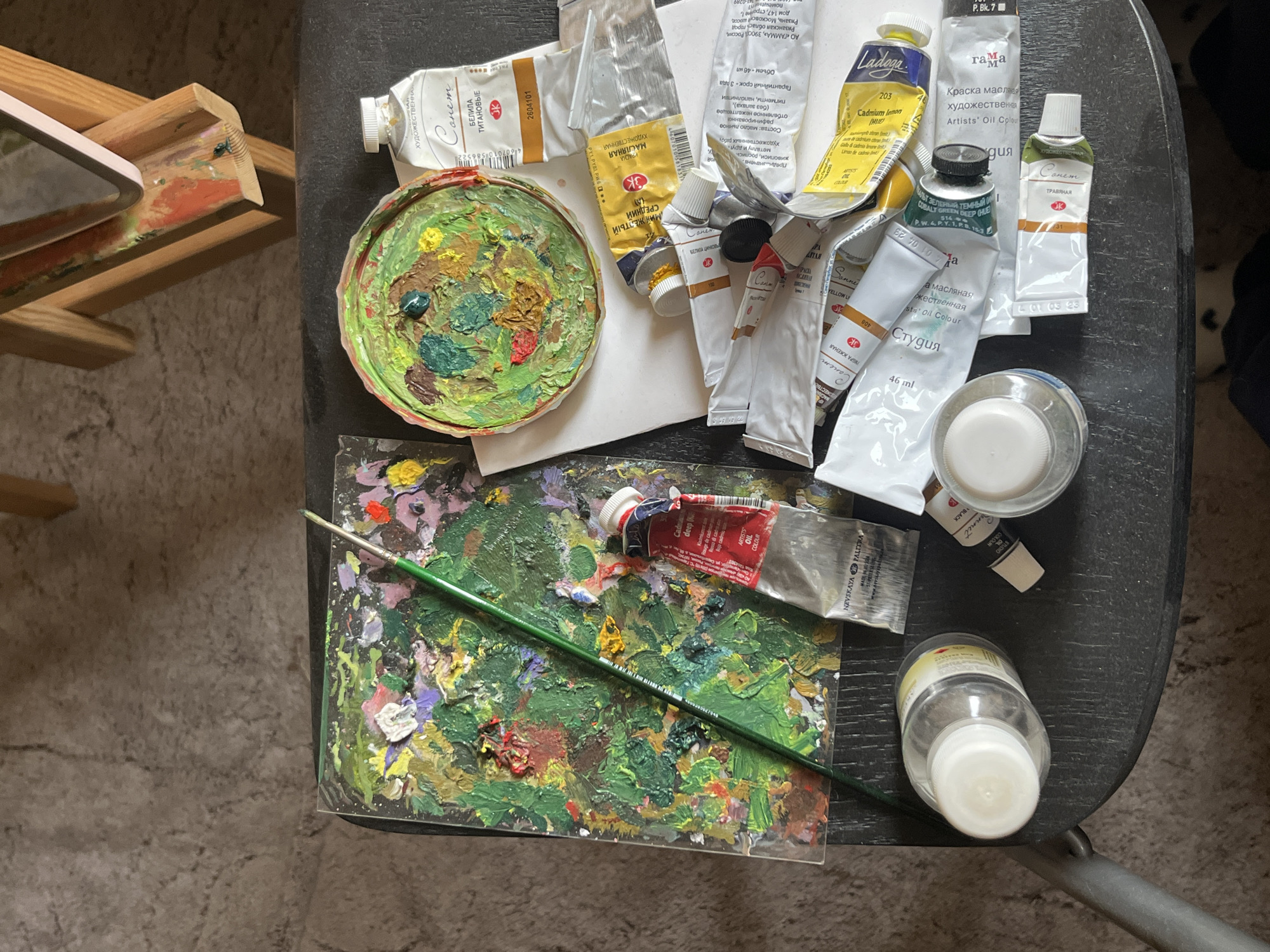 The image shows an overhead view of an artist's workspace. Oil paint tubes and palettes are arranged on a dark, likely metal, folding table. Various tubes of oil paints in shades of red, yellow, green, and white are scattered around the table. Two palettes hold mixed oil paints, one circular and the other rectangular. A green paintbrush lies across the rectangular palette. Two small glass bottles containing liquids, possibly solvents or mediums, are also present. An easel with partially painted surfaces is visible in the upper left corner of the image. The Cyrillic text on the paint tubes suggests the location is Krasnoyarsk, Russia, specifically based on the AO "Gamma" indication that the manufacturer is located in Ryazan, Russia.