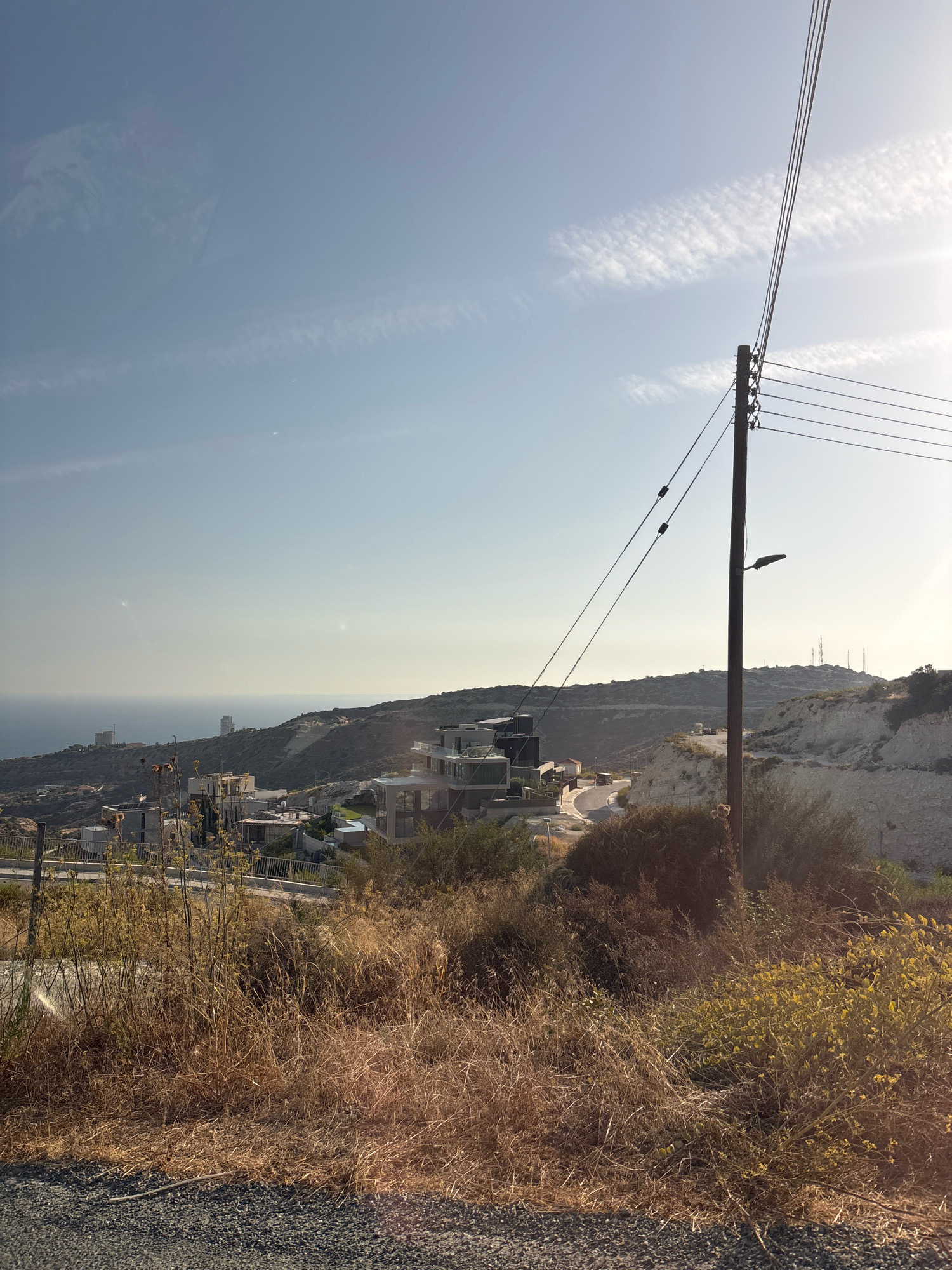 A wide shot captures a sunlit, semi-arid hillside community overlooking the sea in Agios Tychona Community, Cyprus. In the foreground, dry, golden grasses and brush create a naturalistic border along a gravelly path or road. A wooden utility pole with multiple wires stretches diagonally upward into the clear blue sky, which is streaked with faint, wispy white clouds. Beyond the vegetation, a cluster of modern, multi-story buildings with dark facades and large windows is visible on the slope. Further in the distance, the blue expanse of the Mediterranean Sea meets the horizon, with a few taller buildings dotting the coastline. The landscape is characterized by rolling hills with exposed rocky and sandy patches, indicative of dry, Mediterranean terrain. The overall impression is one of a developing or established residential area in a coastal, hilly environment.