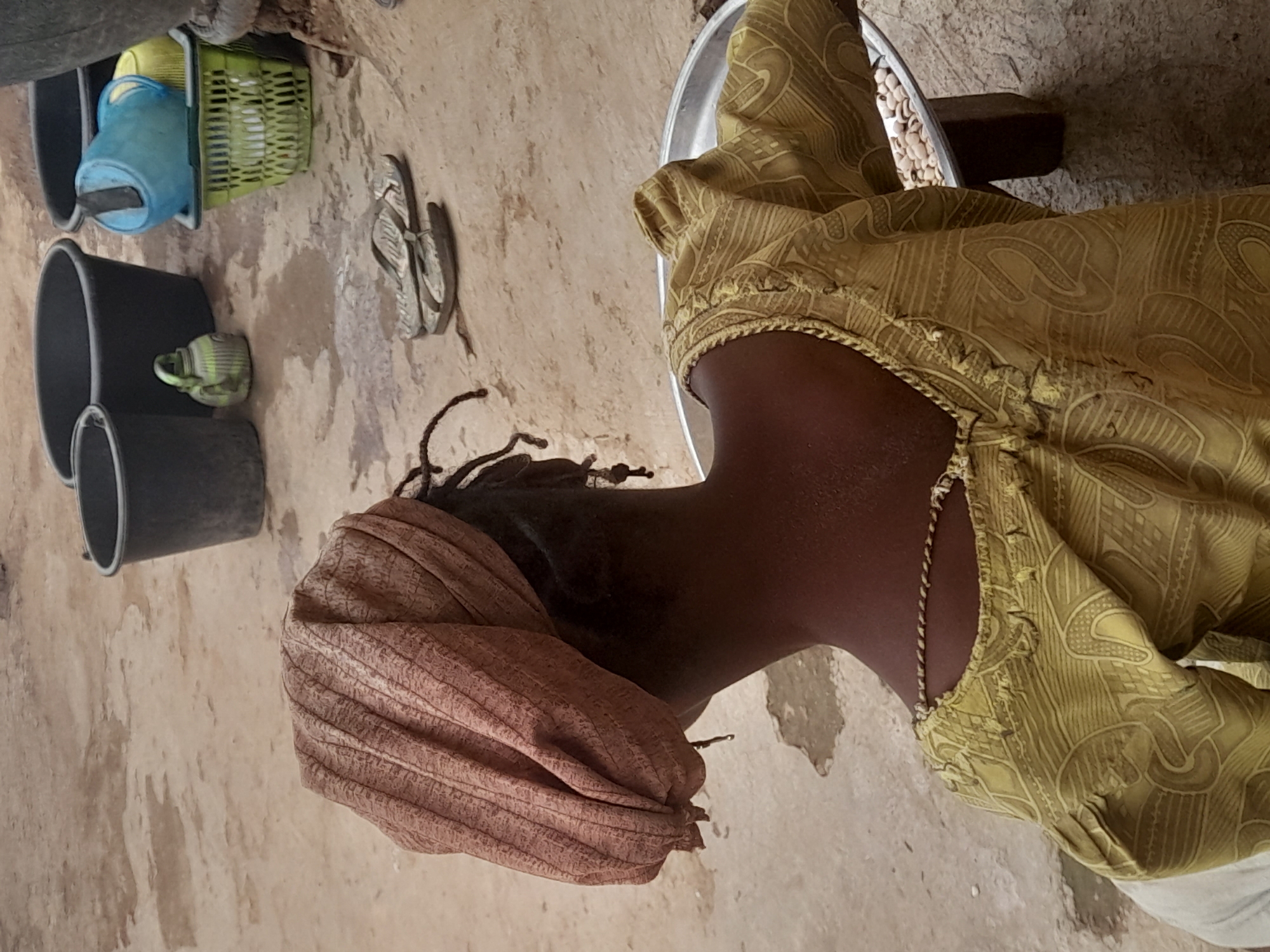 A woman in Akwanga, Nigeria, is seated indoors next to a shallow silver tray filled with what appears to be produce. She is wearing a brown head covering and a yellow patterned top with a chain necklace. To the left of the frame, there are several black buckets and a green plastic basket. A pair of sandals also appears nearby. The room is enclosed with cement walls.
