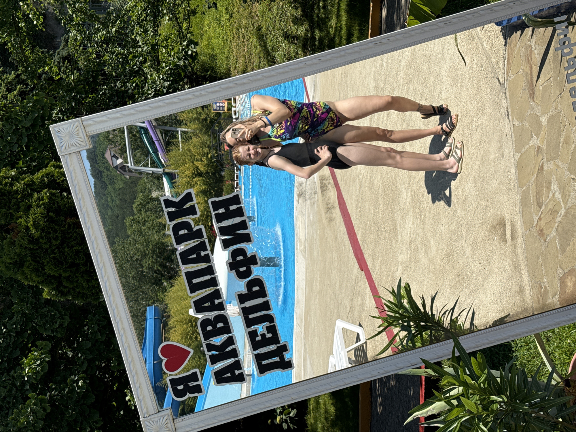 A full-shot, medium-angle mirror reflection shows two young females in swimsuits posing for a selfie at the Dolphin water park in Nebug, Russia. The mirror frame is ornate and white. Text in black, blocky letters is affixed to the mirror, reading "Я ♡ АКВАПАРК ДЕЛЬФИН" which translates to "I ♡ DOLPHIN WATER PARK". Both females are smiling and looking at the camera. The female on the left wears a black one-piece swimsuit and has red hair. The female on the right wears a colorful, patterned one-piece swimsuit and has blonde hair. They are standing on a paved area with a swimming pool behind them. The pool is bright blue and has a fountain in the center. In the background, there are trees and water slides, indicating the location is an outdoor water park. The overall environment is sunny and summery.
