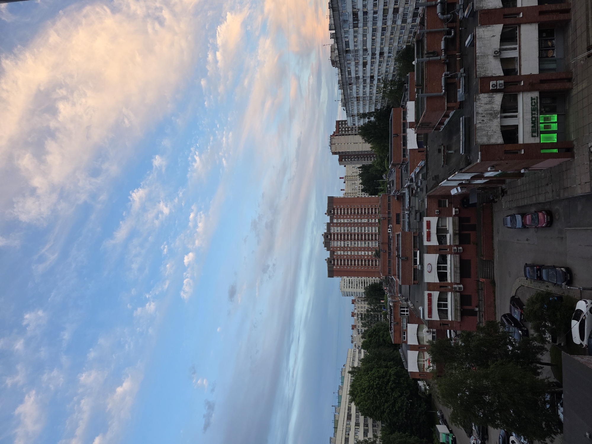 The image captures an urban scene at sunset, characterized by a vibrant sky and dense building development. The upper half of the frame is dominated by a clear blue sky interspersed with various cloud formations; some are wispy and white, while others, particularly those higher up, are tinged with warm orange and pink hues from the setting sun.
Below the horizon, a sprawling cityscape unfolds. On the left and in the background, towering multi-story apartment buildings, predominantly light grey with numerous windows, line the skyline. Moving closer to the foreground and extending across the mid-ground, a series of red brick buildings with white accents and large windows are prominent. These appear to be a mix of residential and commercial structures, with some lower levels seemingly dedicated to businesses.
Green trees with dense foliage are scattered throughout the scene, nestled between buildings and lining streets, adding touches of nature to the urban environment. Several cars are parked along the dark asphalt streets and in adjacent parking areas, suggesting a populated residential or mixed-use neighborhood. The overall lighting is dimming, especially at ground level, indicating evening hours.
A notable detail is a brightly illuminated green sign on one of the red brick buildings, which clearly displays the Cyrillic text "ГЕМОТЕСТ" (Hemotest), identifying it as a medical laboratory. This detail, along with the architectural style common in post-Soviet countries, is consistent with the provided location context of Saint Petersburg, Russia. No people are visible in the image.