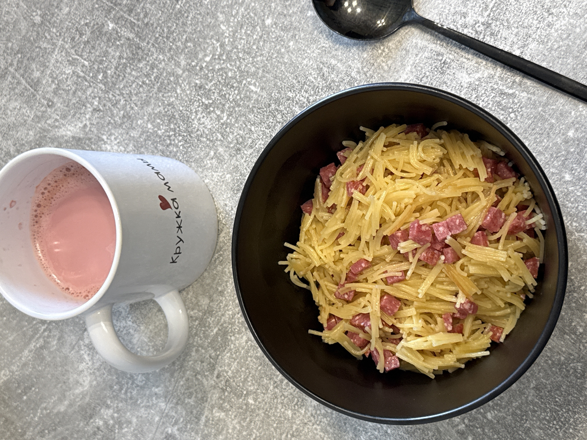 A overhead shot shows a black bowl filled with thin spaghetti and diced salami on a gray, speckled surface. To the left of the bowl is a white mug with a pink beverage and a small red heart and text in Russian, "кружка мамы." To the right of the bowl is a black spoon. The scene is indoors with no indication of a specific city or location.