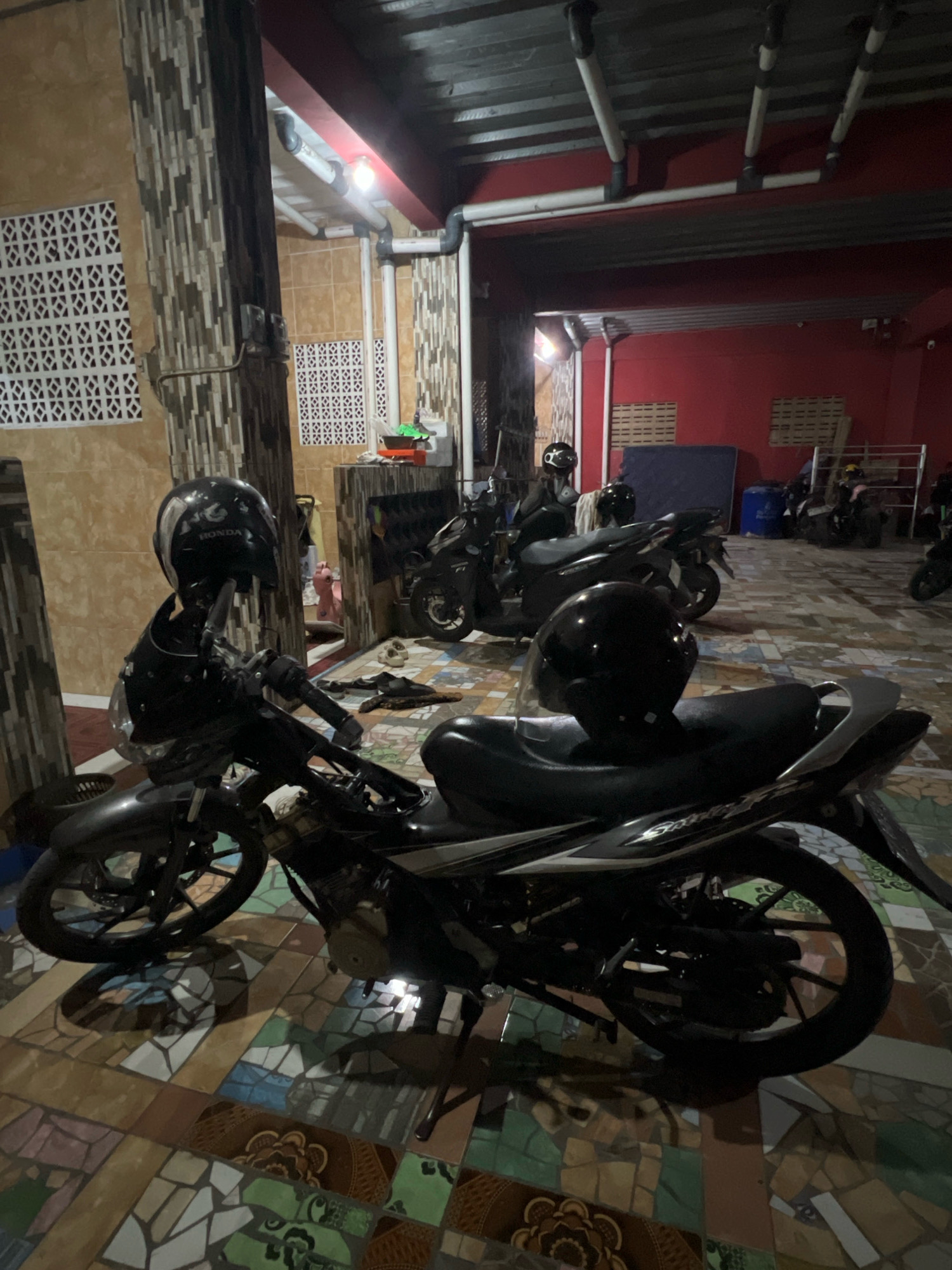 The image depicts a dimly lit outdoor area, likely a covered parking space or a workshop, located in Tlogoadi, Indonesia. In the foreground, two black motorcycles are parked side-by-side. The motorcycle on the left has a black helmet placed on its handlebars. The motorcycle on the right is partially visible, with another black helmet resting on its seat. Both motorcycles appear to be in operational condition.
The environment features a tiled floor with a mosaic pattern composed of various colored rectangular tiles. To the left, a building wall is visible, decorated with a patterned ventilation brickwork. A large, patterned pillar stands next to the wall. In the background, more motorcycles are parked, some with helmets on them. The wall behind the motorcycles is painted red. Various pipes and electrical conduits are visible running along the ceiling. The overall impression is one of a functional, possibly utilitarian, space for storing and maintaining motorcycles.