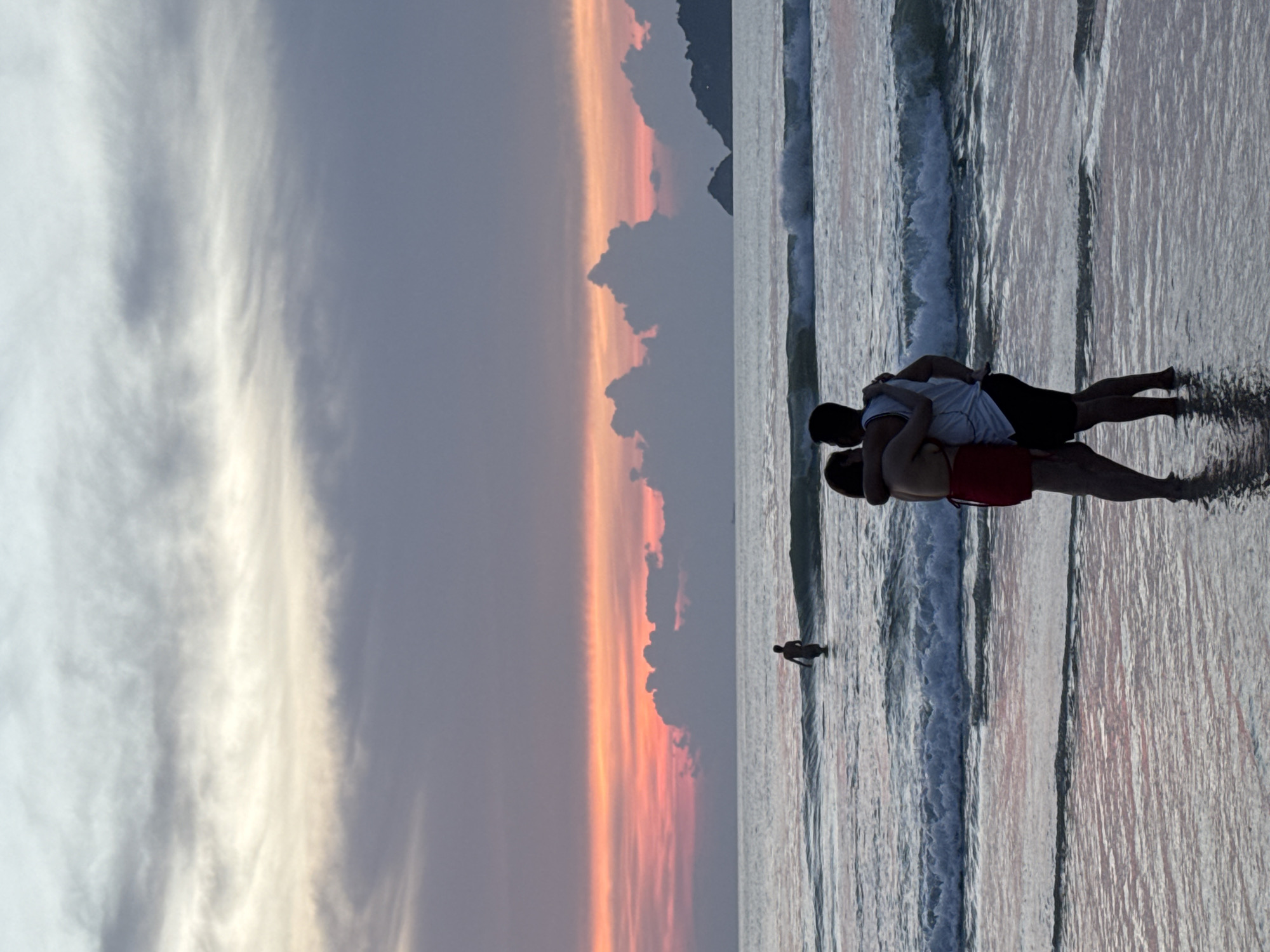 A couple embraces on the shore of Patong Beach in Thailand during a sunset. The man, wearing a white tank top and black shorts, faces away from the camera, his arms around a woman in a red swimsuit. The woman is also facing away, with her arms around the man. Their bodies are partially submerged in shallow water, with gentle waves lapping at their legs. In the background, a single person stands in the ocean, further out from the shore. The sky displays a dramatic sunset, with hues of pink, orange, and purple painted across the clouds. The ocean is a deep blue, with visible waves breaking closer to the shore. In the distance, landmasses with a few scattered clouds are visible on the horizon. The lighting suggests it is either early morning or late evening, with the sun nearing the horizon.