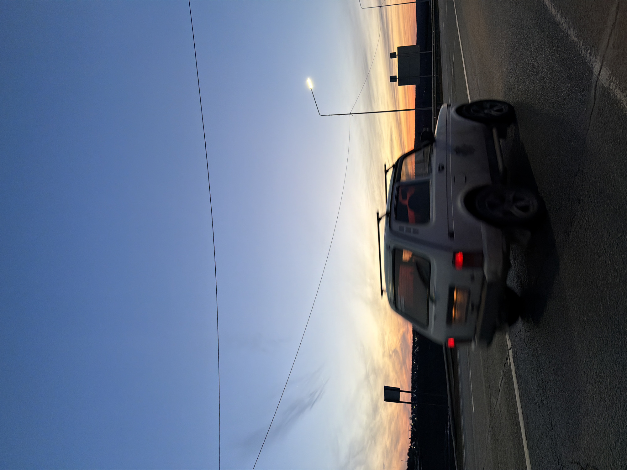The image depicts a gray or white SUV driving on a road at dusk or dawn. The setting appears to be near the edge of a city, with a row of lights suggesting urban development on the horizon. The sky is a gradient of blue to orange and yellow. There are signs present, and a single lamppost is visible. The location is Kuregovo, Russia.