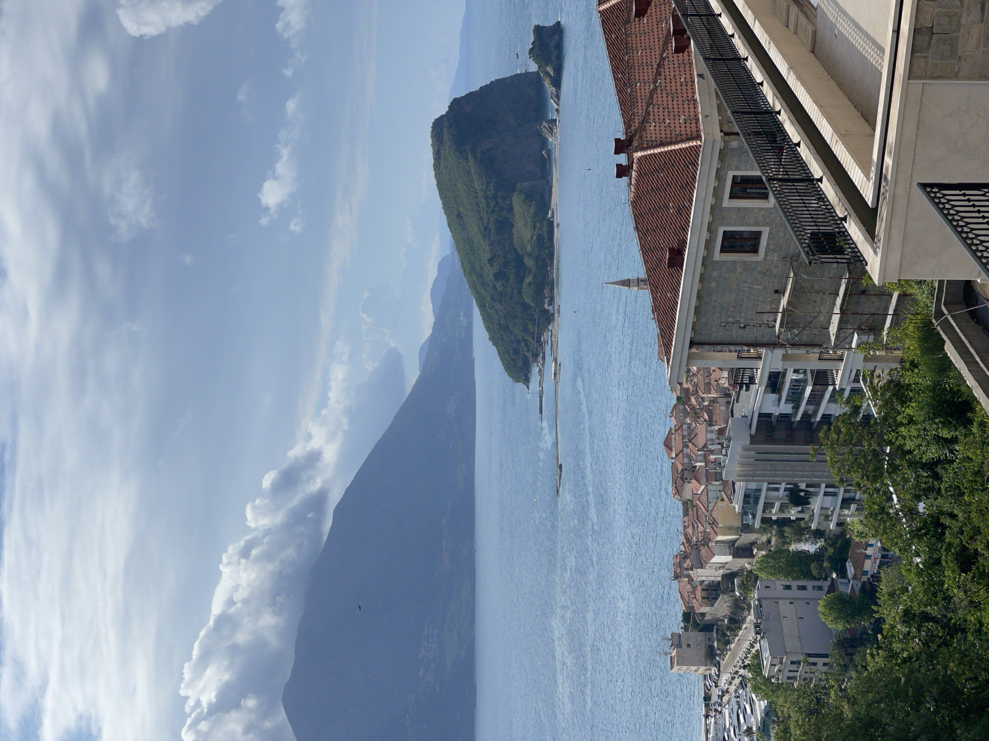 From a high vantage point, the image captures a picturesque coastal view. In the foreground to the right, the architecture of a building, detailed with a tiled roof, windows, and metal balconies, frames the scene. Below, a cluster of buildings with red-tiled roofs and modern structures blend into a cityscape. The Adriatic Sea stretches out, with a densely wooded island in the middle of the frame. To the left, a mountainous landscape recedes into the distance. The sky, filled with clouds, suggests a bright, overcast day. This image likely showcases a panorama from the city of Budva, Montenegro, known for its coastal beauty and historic architecture.