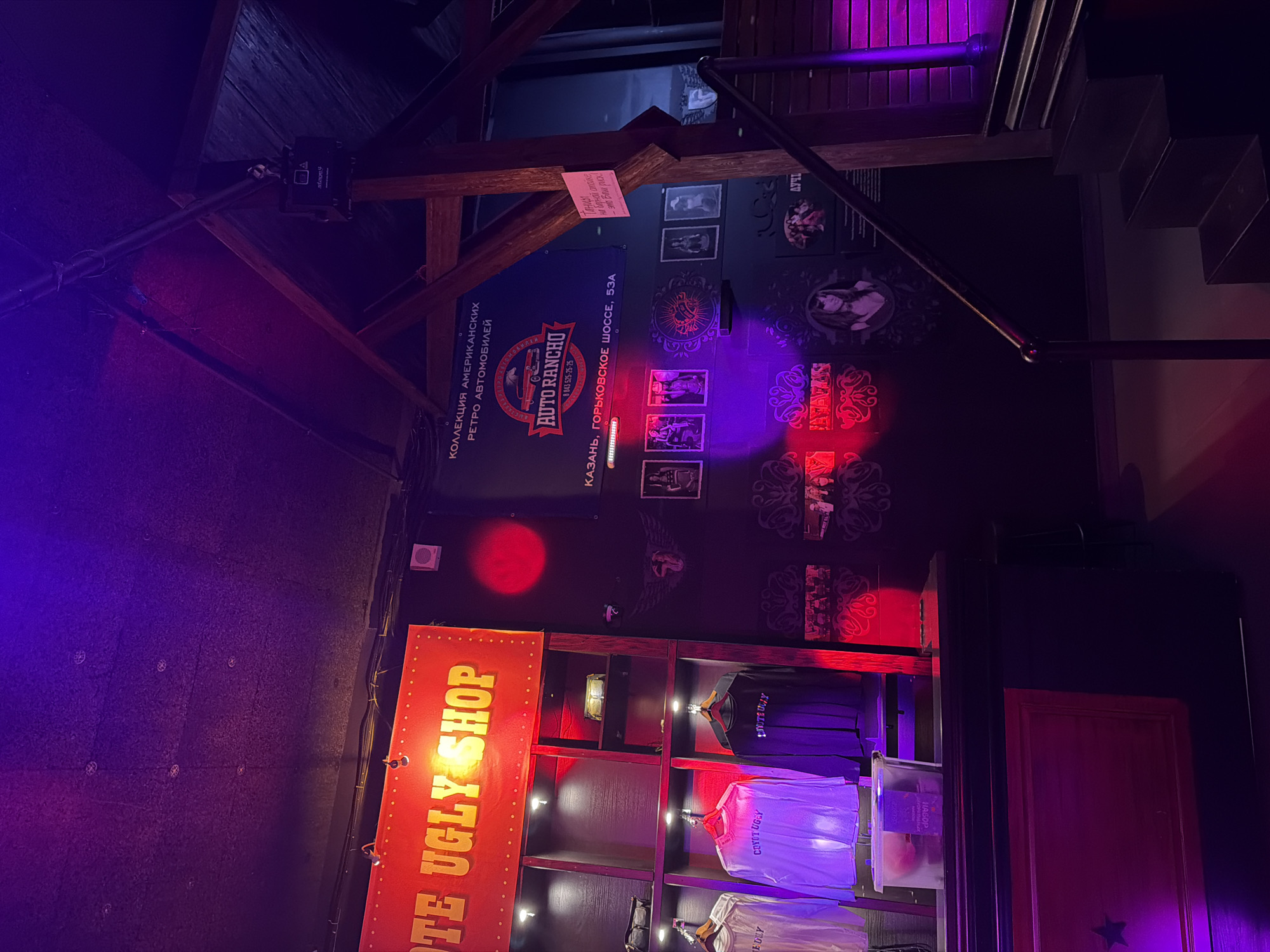 This image captures an interior scene, likely a bar, themed shop, or entertainment venue in Kazan, Russia, as indicated by explicit signage. The setting features a dark, moody ambiance created by strong purple and red spotlights and projected light circles on the walls. Wooden beams are visible overhead, suggesting a rustic or industrial architectural style. A staircase ascends on the right side of the frame.
Key objects include a prominent red sign with illuminated bulbs spelling "TE UGLY SHOP," likely referencing "Coyote Ugly Shop," given the text "COYOT UGEY" and "LAVOTEULLY" visible on T-shirts displayed on shelves beneath it. Another blue banner promotes "КОЛЛЕКЦИЯ АМЕРИКАНСКИХ РЕТРО АВТОМОБИЛЕЙ" (Collection of American Retro Cars) and features the "AUTO RANCHO" logo, along with a phone number (8 843 525-25-25) and address (КАЗАНЬ, ГОРЬКОВСКОЕ ШОССЕ, 53A). A small pink sign on a wooden beam amusingly warns, "Танцы на барной стойке - это Ваш риск!" (Dancing on the bar counter - that's your risk!). The walls are decorated with framed black-and-white pictures, many depicting women in retro style, and intricate circular patterns. Lighting equipment, including a box labeled "stagey," is mounted on the wooden beams.
No people are directly visible, but the decor and signage imply a lively evening or nighttime establishment. The overall atmosphere is dramatic and edgy, characteristic of a nightlife spot or a themed venue. A sign reading "ЛУЧИ" (Rays) is also visible on the far right.