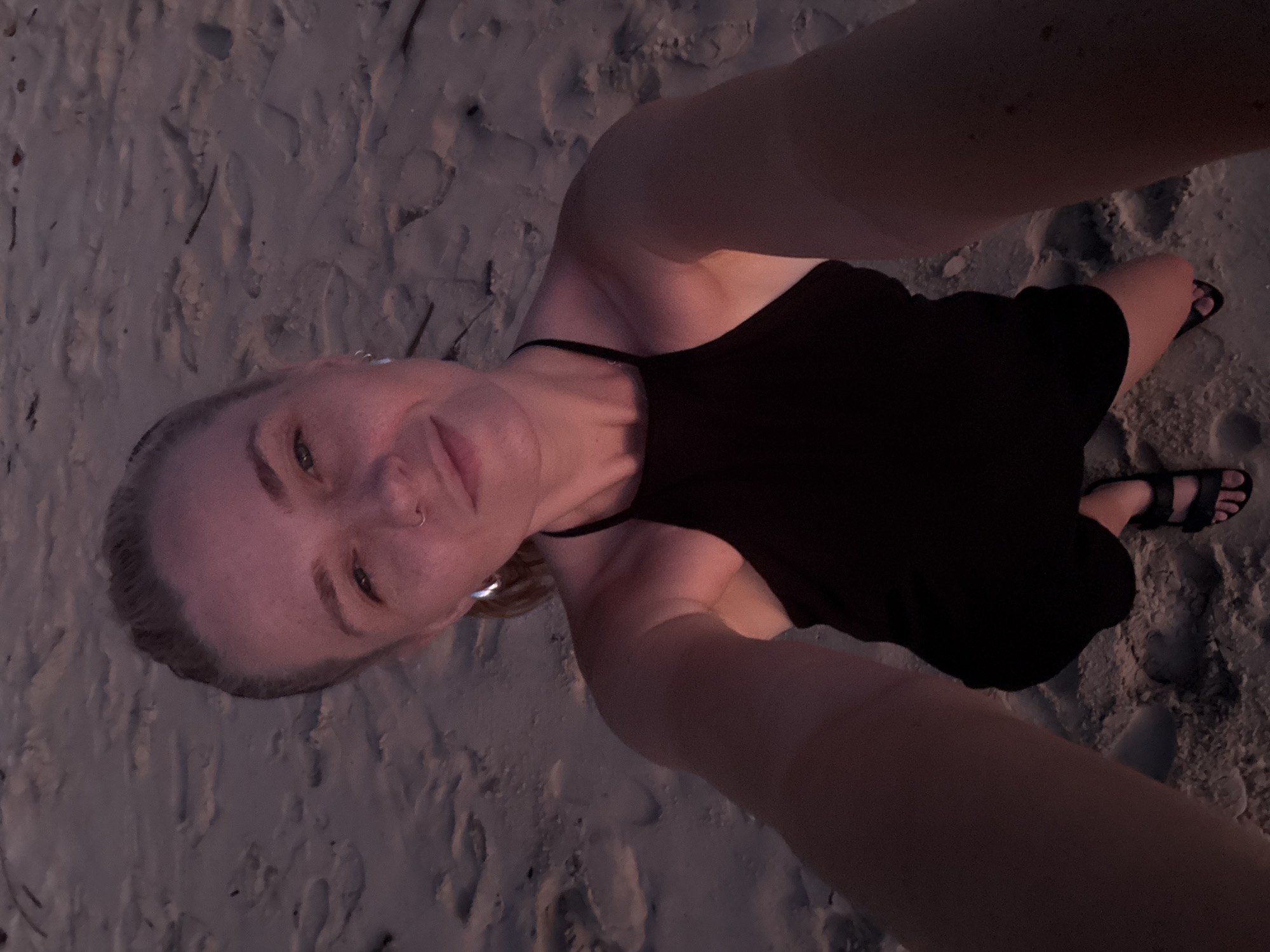 The media file is a selfie taken from a high angle, looking down at a woman lying on a sandy beach. The woman is wearing a black halter dress and black sandals. She has blonde hair and a nose ring. Her eyes are open and she is smiling, appearing relaxed and happy. The sand is textured with footprints and small debris, suggesting it is a natural beach setting. The lighting indicates it is either dusk or dawn, with soft, warm tones illuminating the scene. The woman's arms are visible, one reaching up towards the camera, and the other resting by her side. The overall impression is one of leisure and enjoyment in a natural outdoor environment, likely on a beach in Choeng Thale, Thailand, given the location context.