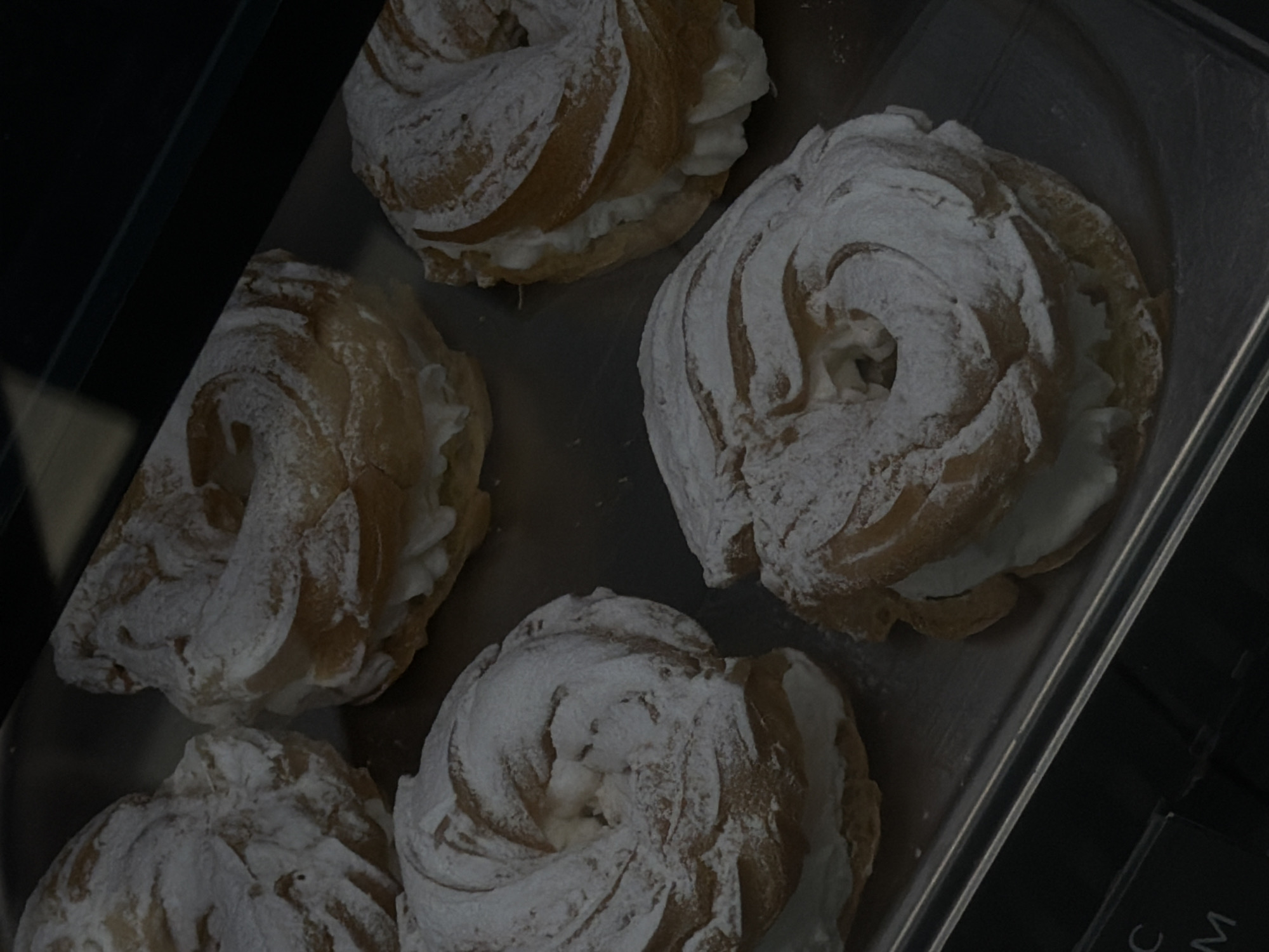 The image shows five pastries, likely cream puffs, arranged on a silver tray. Each pastry has a circular, swirled shape and is heavily dusted with powdered sugar. The pastries appear to be filled with a light-colored cream. The tray is positioned at an angle, and parts of a dark background are visible, possibly indicating a display case or counter. Based on the presence of pastries, the scene is likely in a bakery or cafe. The location in the city of Chaikovsky, Russia is unknown without further evidence.