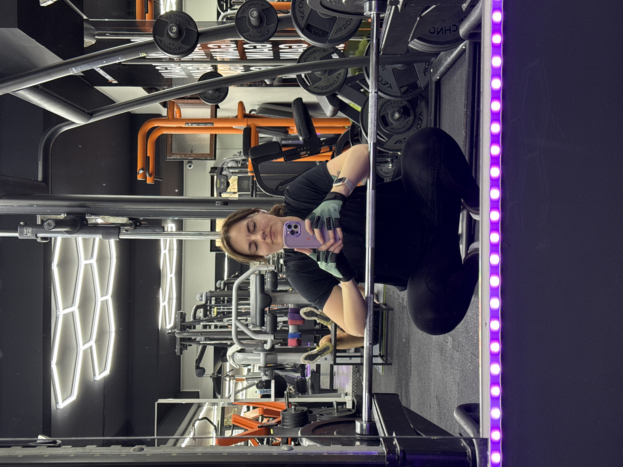 This image shows a person taking a selfie in a gym, possibly in Budva, Montenegro, but this cannot be confirmed without further evidence. The individual is holding a purple smartphone, wearing black clothing, gloves, and a wristwatch. The gym setting includes weightlifting equipment such as barbells, weight plates, and various exercise machines. Hexagonal-shaped neon lights adorn a wall in the background. A column with LED lights is partially visible on the right side of the frame.