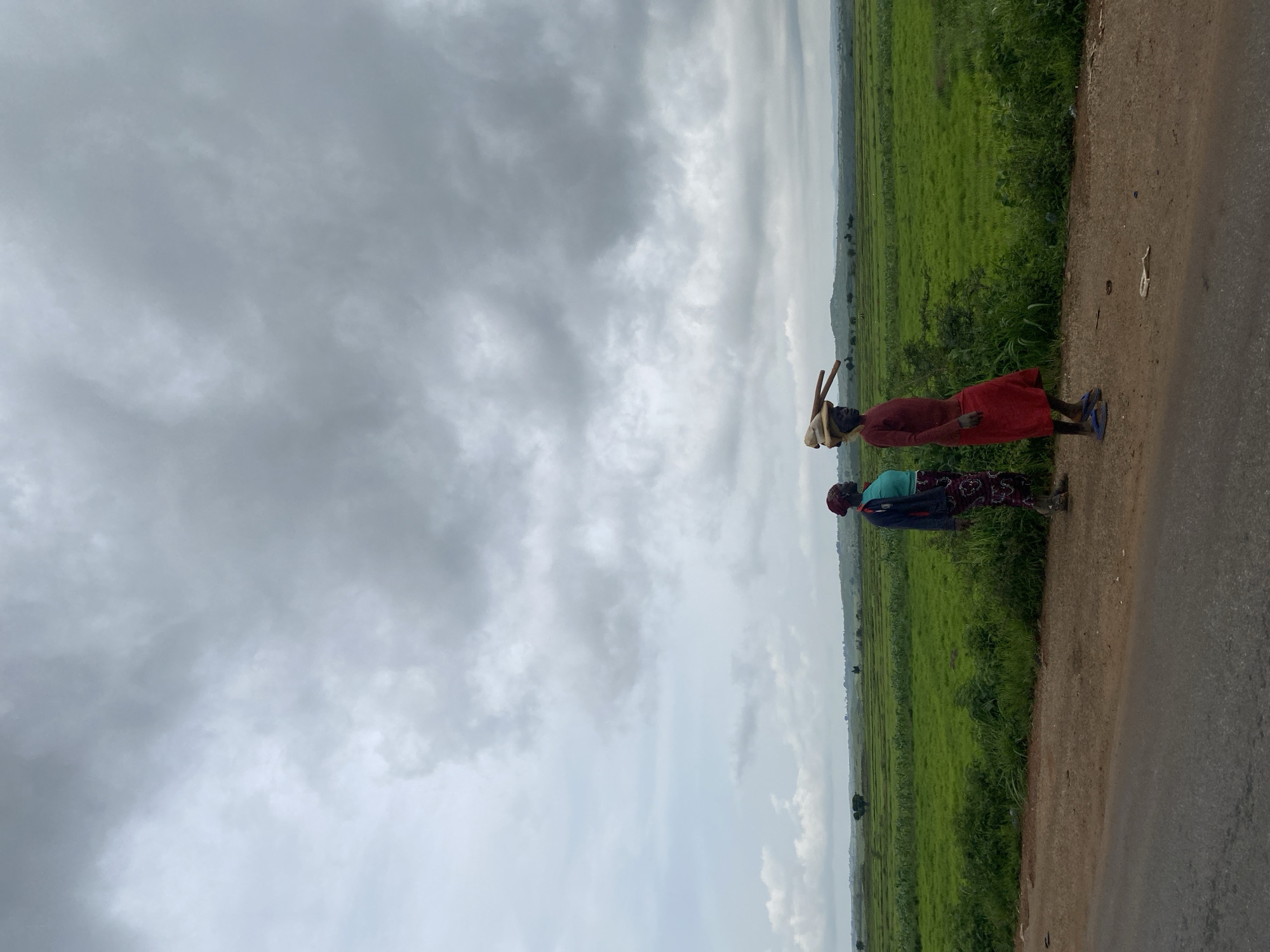 Two individuals stand on a dirt road, facing right. The person in the foreground, wearing a red dress and a headscarf, carries wooden poles balanced on their head. They are barefoot with blue flip-flops visible. To their left, another person wears a turquoise shirt, a patterned skirt, and a dark jacket, also facing right. They stand in a grassy area beside a paved road. Beyond the road, a vast, green, cultivated field stretches to the horizon, meeting a cloudy sky. The scene suggests a rural environment in Makera, Nigeria, possibly during or before a rain event, given the overcast conditions.