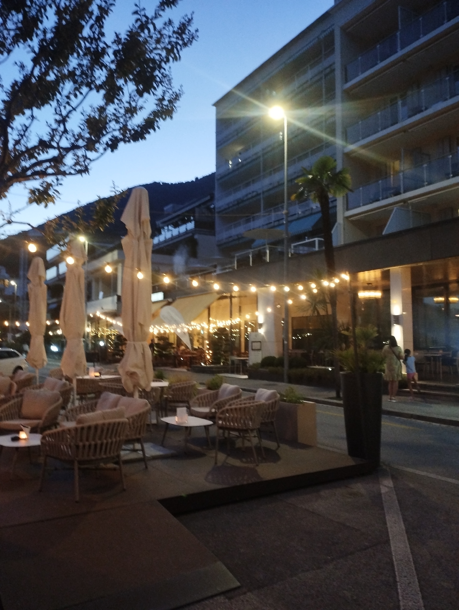 The image depicts an outdoor seating area of a restaurant or cafe on a twilight evening in Muralto, Switzerland. The scene is illuminated by string lights and a streetlamp. In the foreground, there are several tables and chairs made of rattan material, with cushions, set on a dark tiled patio. Some tables have candles or small light fixtures on them.
Behind the seating area, there are tall, closed umbrellas, likely for daytime sun protection. Further back, the facade of a multi-story building is visible, with balconies and windows. A palm tree stands near the building. The street in front of the establishment shows some activity with a few people walking. A light-colored car is parked on the left side of the street. The sky is a deep blue, indicating the transition from day to night. The overall atmosphere is relaxed and inviting, suggesting a pleasant evening for dining or socializing.