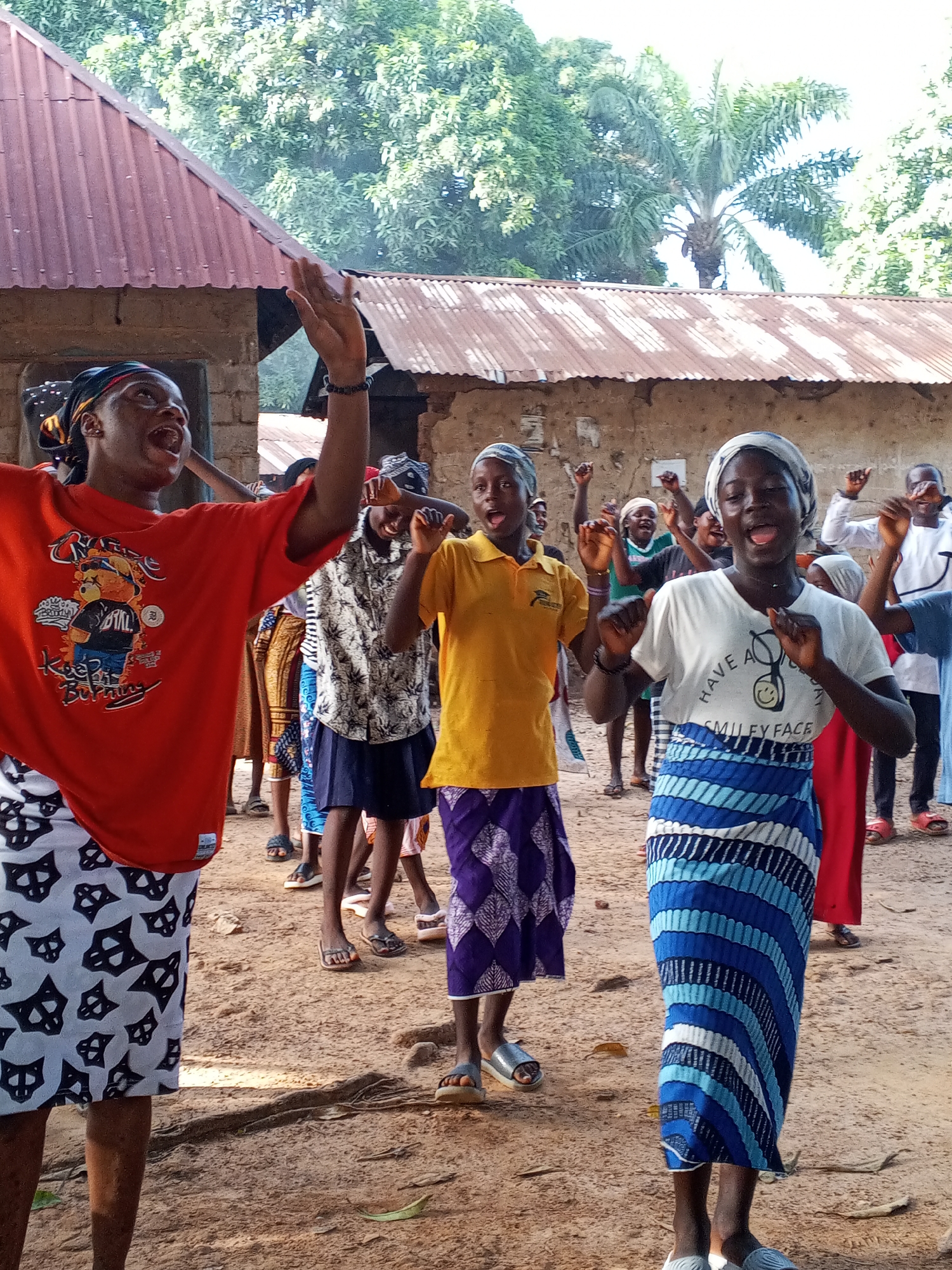 A group of people are gathered outside a building with a corrugated metal roof in Akwanga, Nigeria. The group, comprised of men and women, are singing and raising their arms in the air. They are wearing a mix of modern and traditional clothing, including t-shirts, skirts with patterned fabric, and headscarves. The ground is sandy. There are trees in the background.