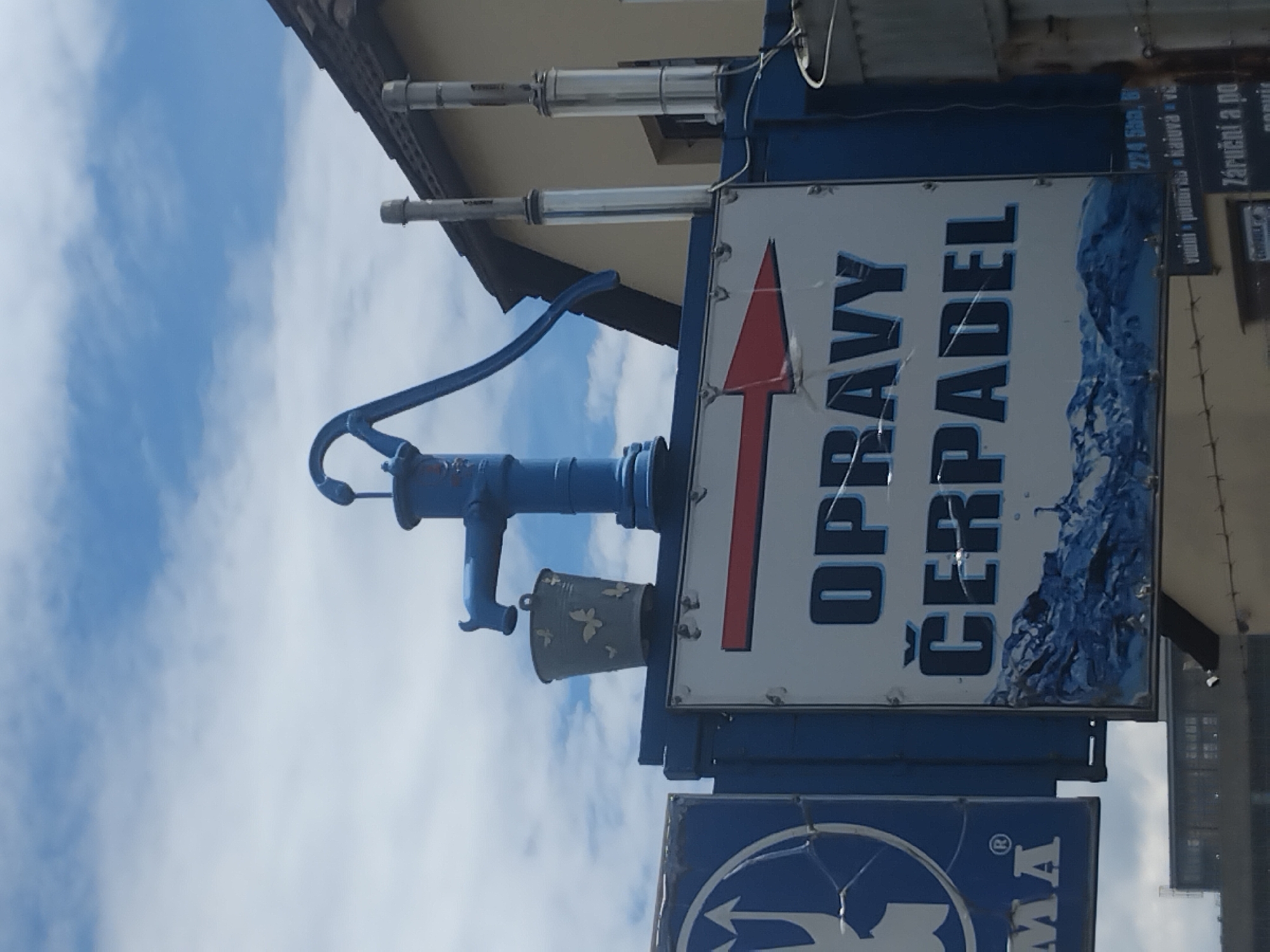 The image shows a blue water pump with a bucket hanging from it, mounted on a sign. The sign, in large blue letters, reads "OPRAVY ČERPADEL", which translates to "PUMP REPAIRS" in English. Below this text, there is a graphic of blue waves, possibly representing water. To the left of the main sign, partially visible, is another blue sign with a white symbol and the letters "MA". The scene is set against a blue sky with scattered white clouds, and a portion of a building with a dark roof is visible in the background. This setup suggests a business that repairs pumps, possibly located in Pilsen, Czechia, given the presence of Czech language text. The overall presentation is a roadside advertisement.