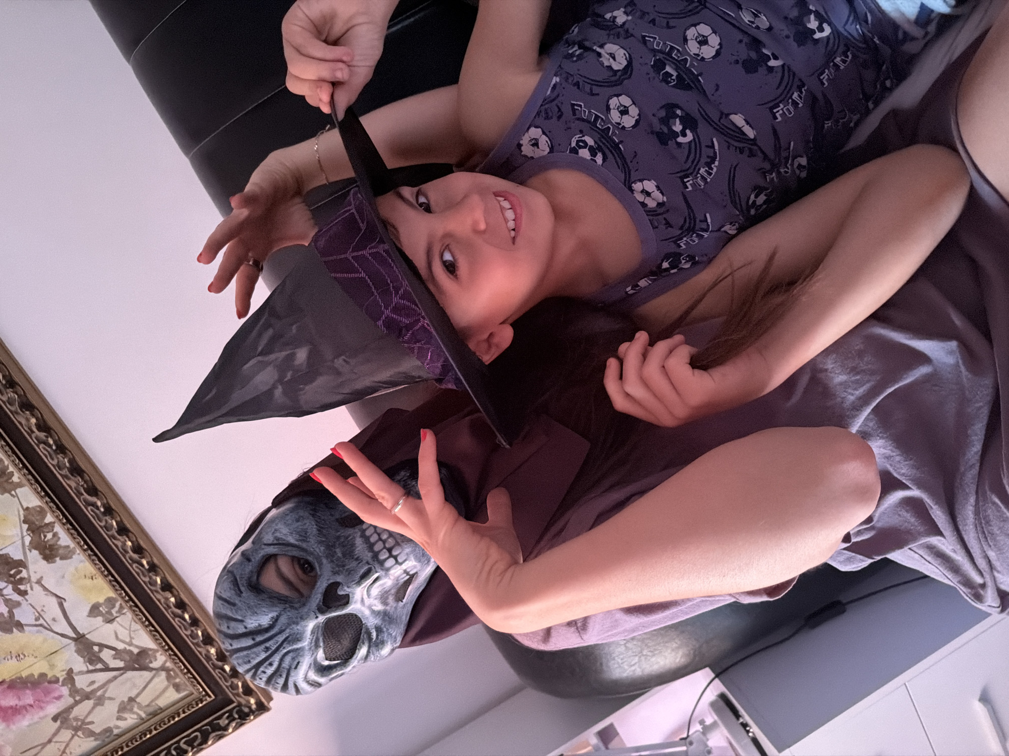 This indoor image, captured in Budva, Montenegro, presents a playful scene featuring two individuals from an overhead perspective.
In the upper portion of the frame, a young child, likely a girl, lies down and smiles directly at the camera with a bright, joyful expression. She has dark hair and wears a grey or light purple tank top featuring "FUTSAL" text repeated alongside stylized soccer ball graphics. Her left hand is raised, seemingly interacting with the witch's hat, while a thin bracelet adorns her wrist.
Positioned below the child is an adult, partially obscured by a costume. The adult wears a grey skull mask with blue-grey accents, completely covering their face. A black, pointed witch's hat, adorned with a purple patterned band, rests on their head. One of the adult's hands is visible near the mask, showcasing bright red nail polish and a silver-colored ring on the ring finger. This hand appears to be resting or gesturing. The adult is dressed in a dark top, consistent with the costume theme.
The setting appears to be a casual indoor environment, possibly a living room or bedroom. Behind the child is a dark, possibly black, sofa or bed. To the left of the adult's mask, an ornate gold-framed picture with a delicate floral or branch design is partially visible. In the lower right corner, a light-colored surface, such as a table or desk, and part of a dark electronic device can be seen. The overall atmosphere of the image is cheerful and fun, suggesting a moment of dress-up or playful interaction. The "FUTSAL" text on the child's shirt is clearly legible.