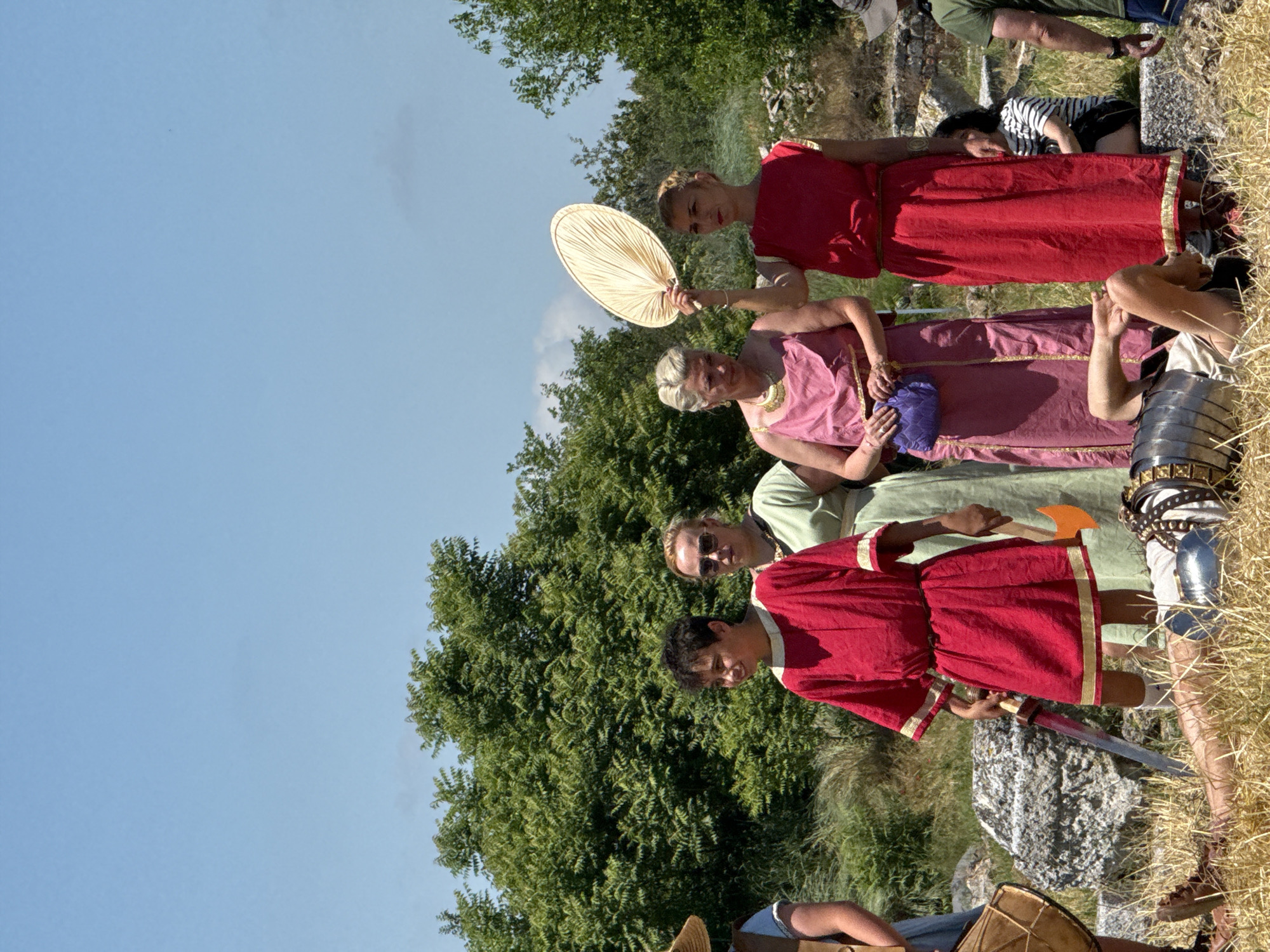 The image captures an outdoor historical reenactment scene in Gigen, Bulgaria, under a clear, bright blue sky with scattered clouds. In the foreground, a person lies on their back in dry, yellowish grass, wearing a light-colored tunic and Roman-style segmented armor (lorica segmentata), portraying a fallen figure. Standing over them to the left is a young individual in a red knee-length tunic with gold trim, holding a silver sword in their right hand and a small orange axe in their left hand. To the immediate left, a partial view of a person in a straw hat holding a large, round, leather-bound drum is visible.
Behind these foreground figures, four women stand in a line. From left to right: one woman with short blonde hair and sunglasses wears an olive-green dress and a dark necklace; next to her, a woman with blonde hair in an updo wears a pink sleeveless dress with gold trim, holding a purple clutch bag; to her right, another woman with blonde hair in a bun wears a red sleeveless dress with gold accents, holding a light-colored pleated fan aloft in her right hand. Further to the right, a person in a green t-shirt is partially visible. In the background, a person wearing a striped shirt and dark shorts sits on a stone block, observing the scene.
The environment features dense green deciduous trees across the midground and background, with large grey stones and remnants of what appear to be ancient ruins scattered on the ground. The ground is covered with dry grass and dirt.