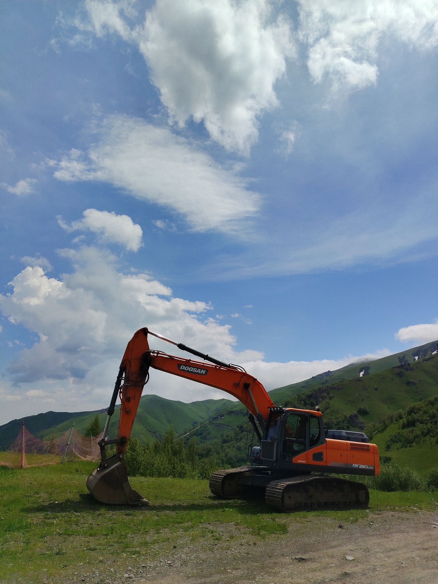 An orange Doosan excavator, model DX 260LCA, is parked on a grassy slope in Bakuriani, Georgia, with a mountainous landscape and blue sky with scattered clouds in the background. The excavator's bucket is lowered to the ground. A pink safety net is visible on the left side of the frame. The terrain is uneven, with both grass and gravel visible. There are no people or animals in the image.