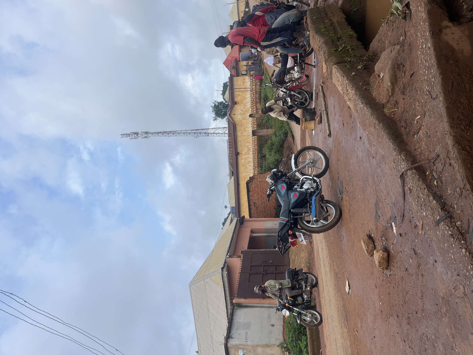 The image depicts a scene in Gembu, Nigeria, characterized by a collection of motorcycles and people gathered on a dirt road. The atmosphere appears to be a typical day, with overcast skies and a generally muted color palette dominated by browns and grays, suggesting a cloudy or possibly rainy day.
Several motorcycles are parked or in use. In the foreground, a person is seated on a black motorcycle, wearing a light-colored shirt and a traditional cap. Further up the road, another motorcycle, this one black with blue accents and a red emblem, is partially visible, with a rider also on it. In the background, a group of people are clustered around motorcycles, some standing and others seated. They are dressed in casual clothing, with one individual in a red shirt and another in a striped garment.
The setting is rural or semi-urban, with buildings visible in the background. One building has a corrugated metal roof and light-colored walls. Another structure further back appears to be made of brick. A prominent feature in the distance is a tall telecommunications tower, indicating a degree of modern infrastructure within the locale. The road itself is unpaved, with areas of mud and scattered debris, common in many Nigerian towns and villages. The overall impression is one of everyday life, with motorcycles serving as a primary mode of transportation and a social gathering point. No discernible emotions are evident from the individuals depicted, as the focus is on the static scene. There is no visible text that is clear enough to be read.