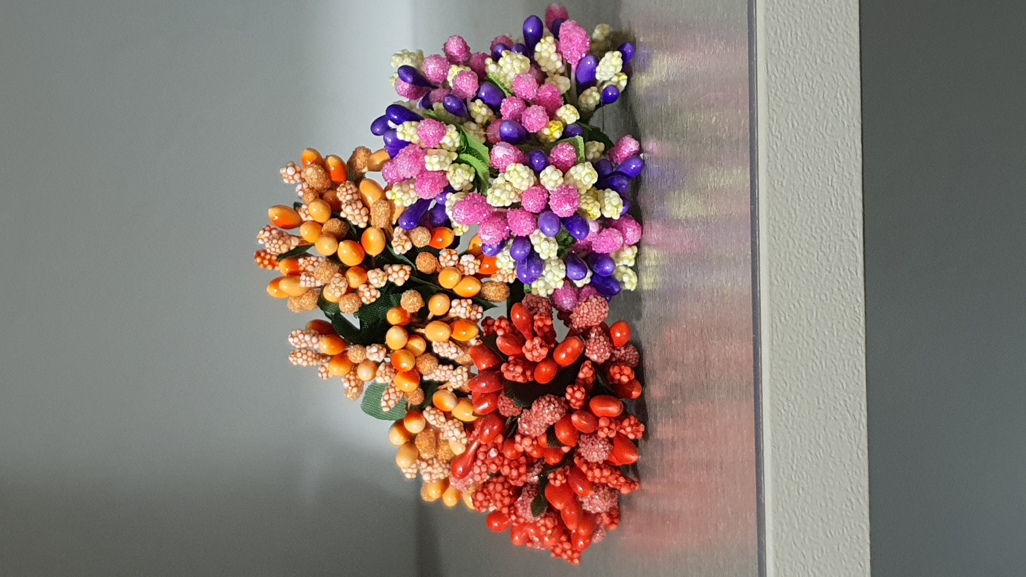 The image shows a close-up, eye-level shot of three small bunches of artificial floral embellishments. The subjects are arranged in a cluster on a metallic, reflective surface, likely a shelf or table. The background is a plain, gray, vertical surface with a subtle gradient, suggesting a studio or indoor environment. The floral embellishments are made of small, bead-like elements clustered together on short stems, forming rounded shapes. They are colored in distinct groups: one bunch is bright red, another is orange and light brown, and the third is a mix of pink, purple, and white. There are no people, animals, or any indication of a specific location in Krasnoyarsk, Russia, or any other city. The lighting appears to be artificial and comes from the top, casting soft shadows. The overall impression is one of a still life arrangement of craft supplies.