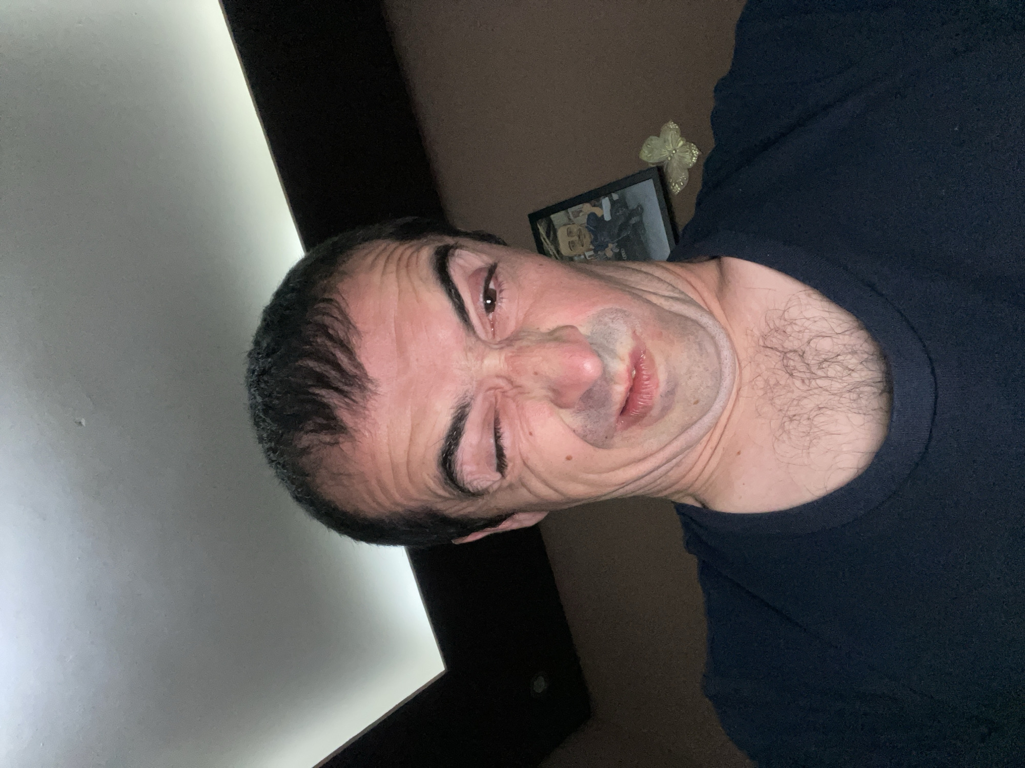 A man with dark hair and a dark t-shirt is making a facial expression with his eyes squinted and nose wrinkled. The background includes a white wall, a dark wall, and a framed picture. The location cannot be determined from the image.