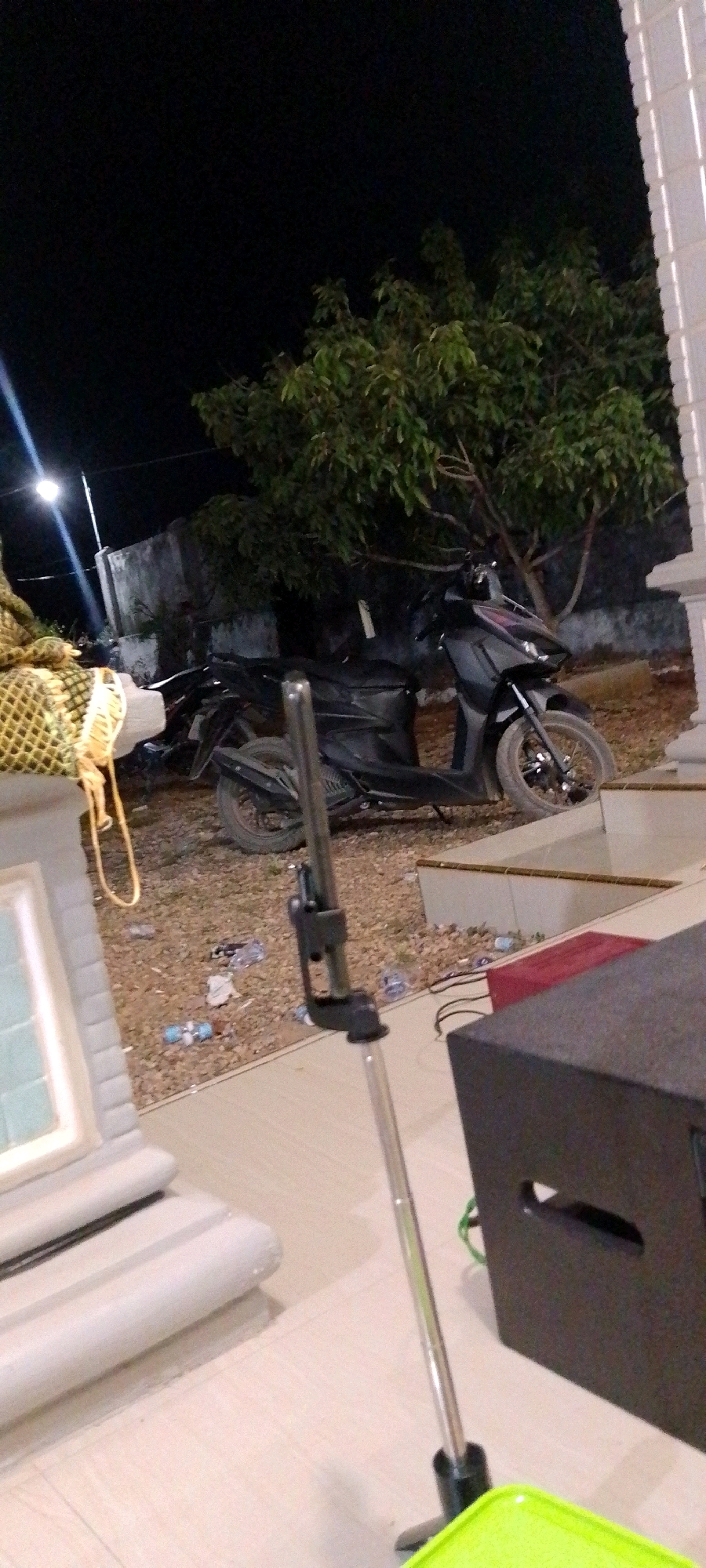 A black motorcycle is parked on a gravelly area next to a white building with white vertical panels. Trees with green leaves are behind the motorcycle. A thin metal pole with a black attachment is in the foreground, and a black speaker is to the right of the pole. A light pole with a light at the top is visible in the background to the left of the motorcycle. Scattered on the ground are plastic bottles and other debris. The scene appears to be outdoors at night.